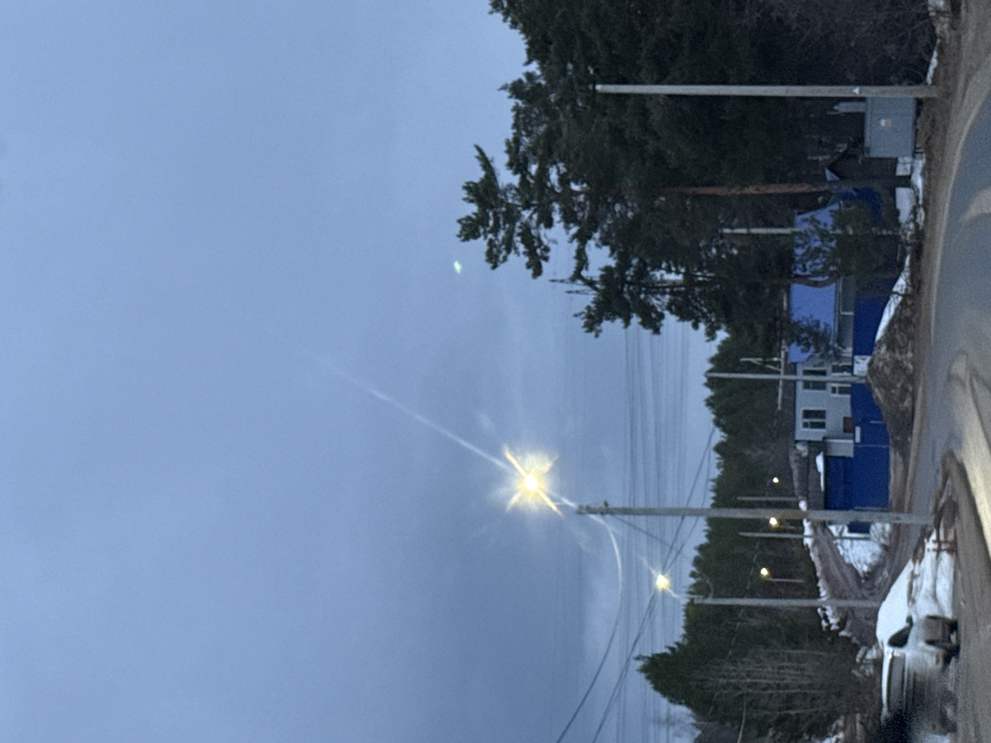 The image shows a street scene in what is assumed to be the city of Chaikovsky, Russia. The sky is overcast, suggesting either early morning or late afternoon. There are several streetlights illuminated, indicating low light conditions. Power lines are visible. Along the roadside, there is a mixture of coniferous trees and buildings, some of which are blue. A vehicle is parked on the side of the road, partially covered with snow. Patches of snow are also visible on the ground.
