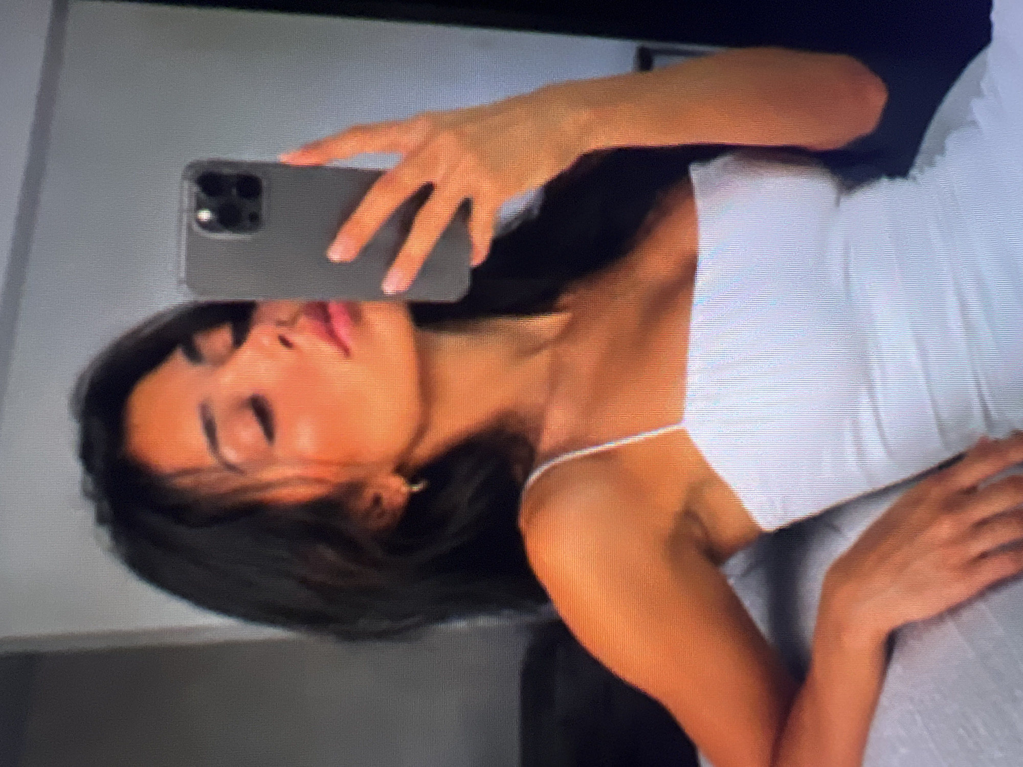 A young woman with dark hair takes a selfie in a mirror, holding a gray smartphone. She is wearing a white spaghetti strap dress and has an earring in her left ear. She is indoors, and the background is blurred, with a dark sofa visible to her left.