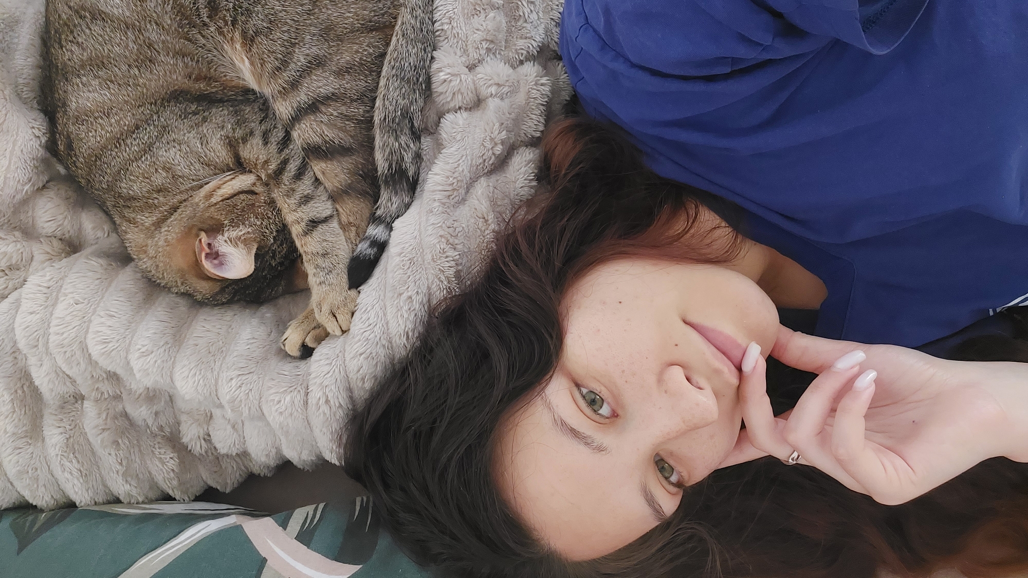 The image depicts a young adult female and a sleeping tabby cat lying together on a soft, textured, light grey blanket, located in the city of Shumen, Bulgaria. The female is positioned in the foreground, looking directly at the camera with a subtle smile. She has long, wavy dark brown hair, green eyes, and visible freckles across her nose and cheeks. She wears a plain navy blue t-shirt. Her left hand, adorned with a silver ring on the ring finger and neatly manicured nails with light-colored polish, rests gently against her chin. Above and to the right of the female, a brown and grey striped tabby cat is curled up and appears to be asleep, with its head tucked under its body and paws. Its striped tail is visible. To the far left of the frame, a segment of a green fabric item with a lighter abstract pattern is visible, likely a pillow or cushion.