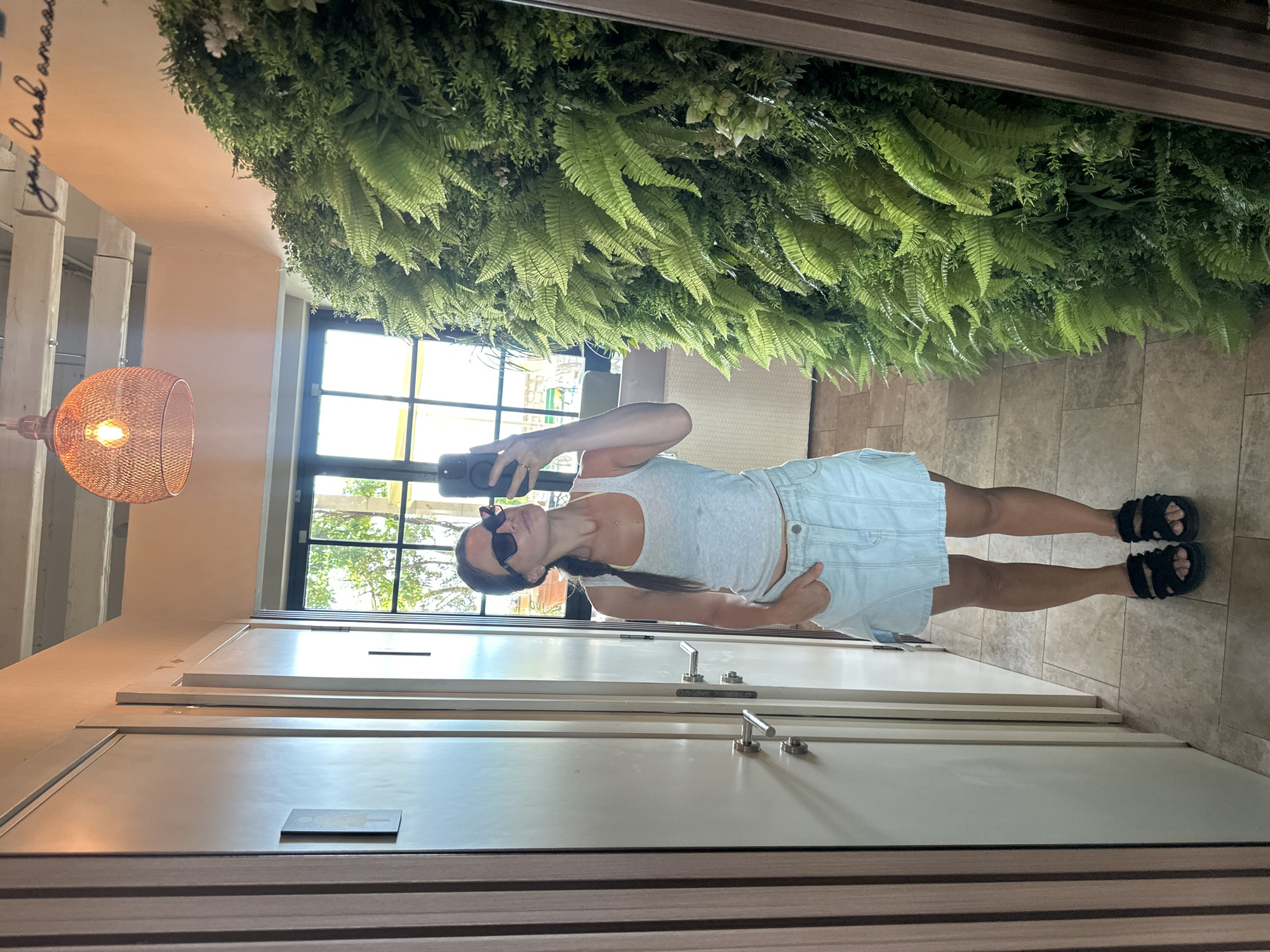 A woman stands in front of a full-length mirror, taking a selfie. She is wearing a light gray tank top, a light blue denim skirt, and black platform sandals. Her hair is pulled back into a braid, and she is wearing sunglasses. To her right, a large wall of lush green ferns and other plants fills the frame, with a glimpse of a window and an outdoor scene beyond. To her left, there are two closed doors with modern silver handles. The ceiling has exposed beams and a large, decorative light fixture. The floor is tiled with beige squares. Above the green wall, a motivational quote is handwritten on the peach-colored wall in black cursive script. The overall impression is of a stylish, modern interior, possibly a changing room or a private area in a boutique or hotel. The location is Bečići, Montenegro.