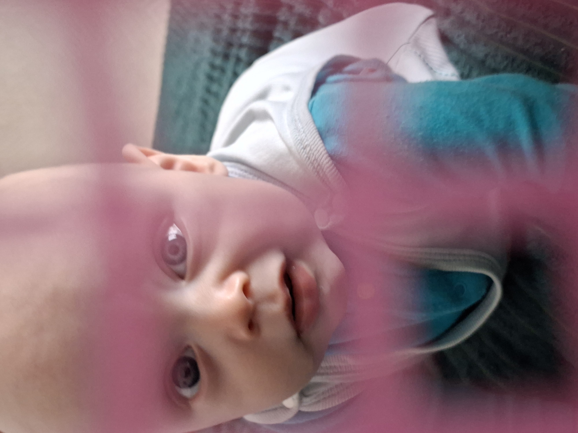 A baby lies on a dark green textured surface with a white and blue shirt. A pink, out-of-focus object is between the camera and the baby. The location is unknown.