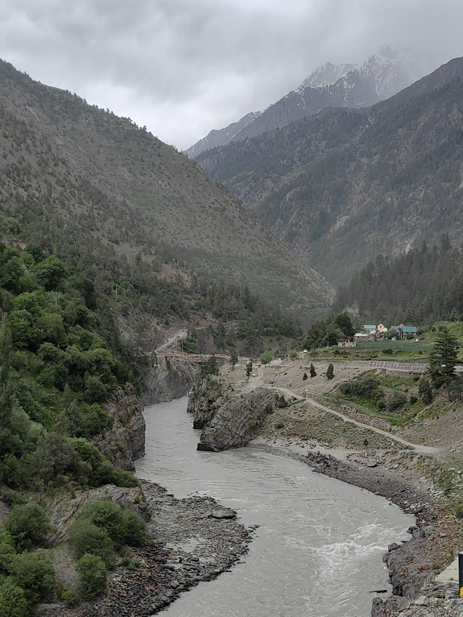 The image depicts a vast, mountainous landscape with a wide, turbulent river flowing through a valley. Steep, rocky slopes covered in sparse vegetation, primarily trees and shrubs, flank the river. The sky is overcast, contributing to a muted, somewhat somber atmosphere. In the distance, snow-capped mountains are visible, suggesting a high-altitude location. A road is visible winding along one of the slopes, and a small cluster of buildings, possibly a village or settlement, is nestled in the valley. The image does not contain any people or specific actions. The location is identified as Arat, India, a region known for its dramatic mountain scenery and river valleys.