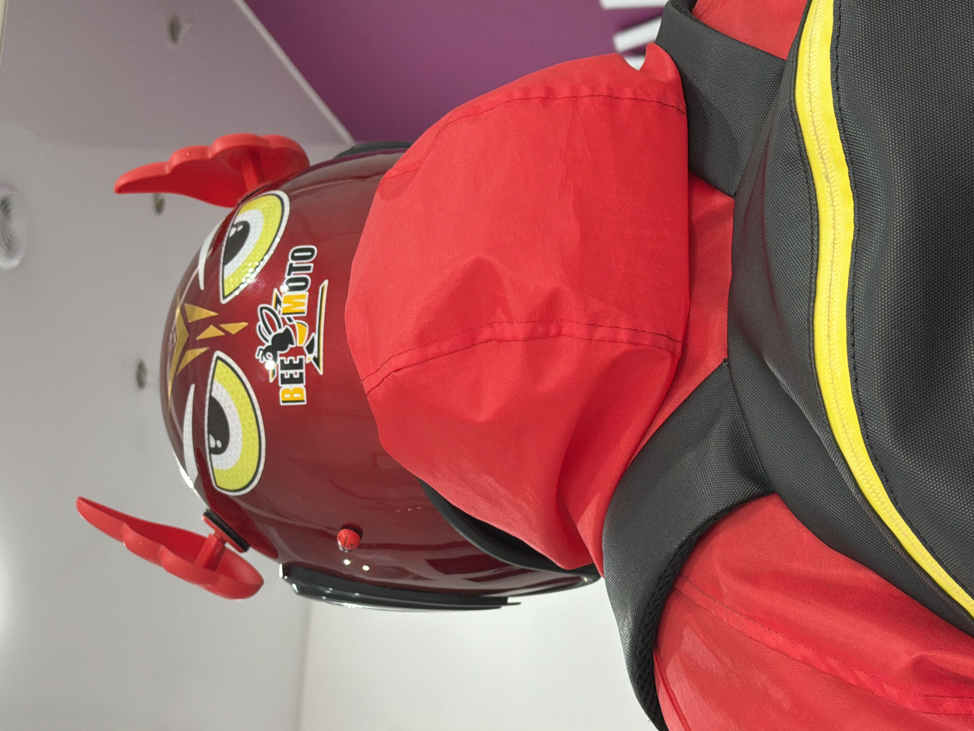 A close-up shot, taken from a low angle, reveals a red motorcycle helmet with attached red wing-like elements, positioned adjacent to a black and red garment. The helmet features cartoonish eye graphics and a logo with the words "BEE MOTO". The garment has black straps with a yellow border. The background shows a neutral-colored wall meeting a colored surface in shades of pink and violet. The location cannot be determined from the image.