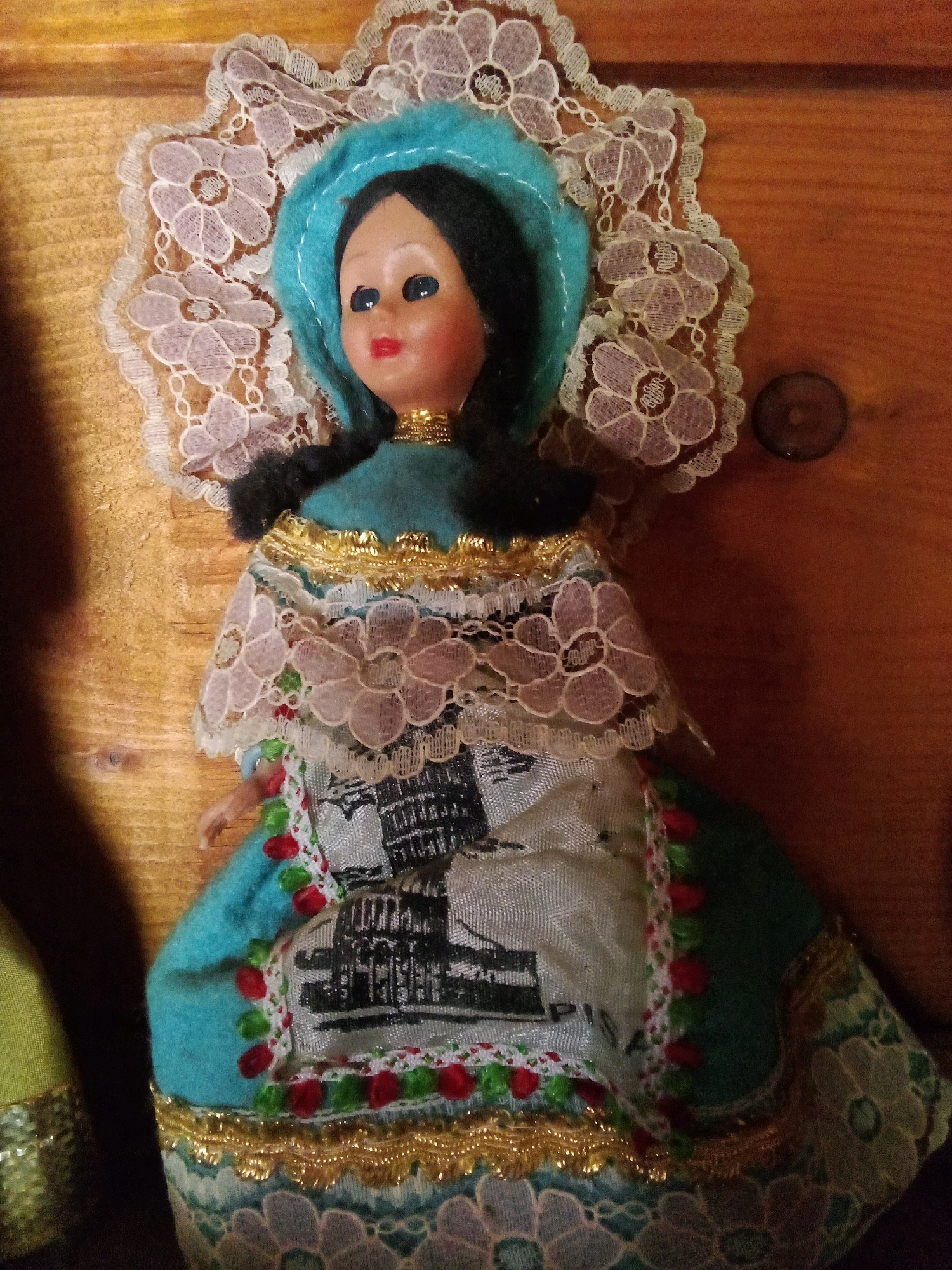 A vintage doll, dressed in a traditional Italian costume, is positioned against a wooden background. The doll features a light blue felt hat, dark braided hair, and painted facial features. Her elaborate dress is adorned with white lace, gold trim, and a printed depiction of the Leaning Tower of Pisa. The background is a warm-toned wooden surface with visible grain and a single dark circular object, possibly a coin, to the right of the doll. There is no indication or context within the image that places it in Santomenna, Italy.