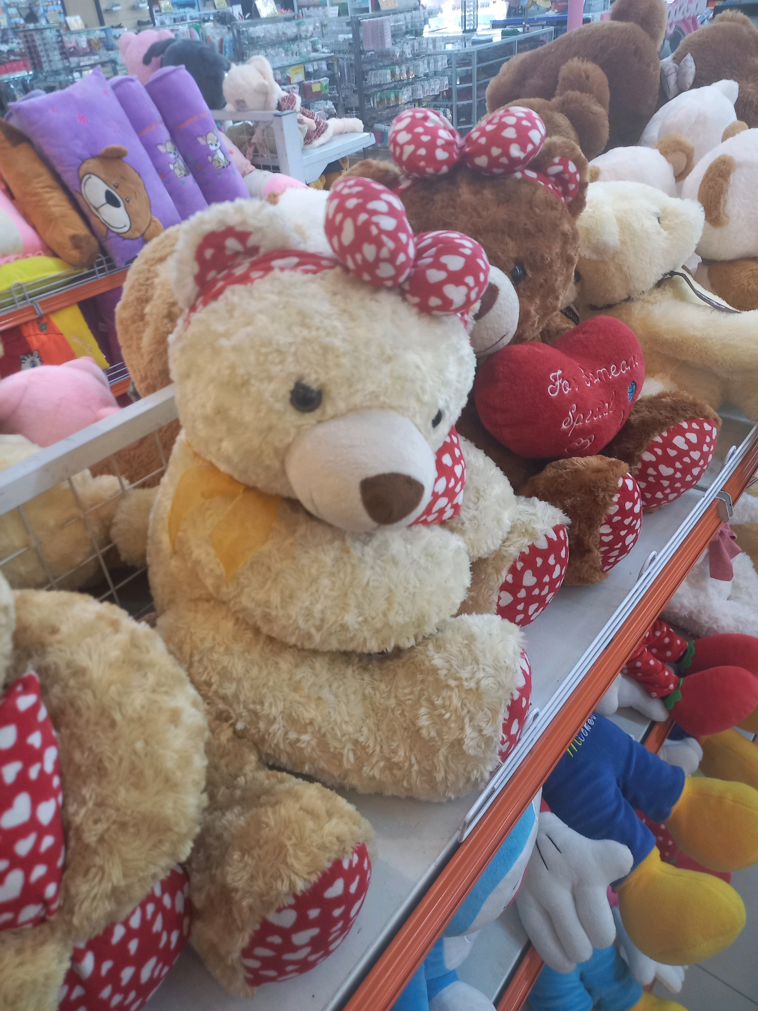 The image shows a close-up of a shelf filled with stuffed toys in what appears to be a retail store. The primary focus is on several large teddy bears, with cream and brown fur, adorned with red and white heart-patterned bows and fabric accents. One cream-colored bear in the foreground is particularly prominent, with a yellow ribbon tied around its neck. Behind it, a brown bear with red bows is visible, along with other teddy bears and pillows of various sizes and colors, including purple pillows with teddy bear faces. Further back, more stuffed animals and merchandise can be seen on shelves, suggesting a toy store or a section of a larger store. The overall impression is one of a brightly lit retail environment showcasing a collection of plush toys. The location is identified as Tunggorono, Indonesia.