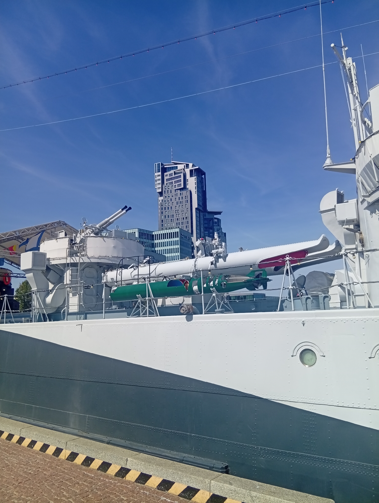 A medium shot, taken from a slightly low angle, captures a sunny day with a bright blue sky and a few wispy clouds. The foreground is dominated by the side of a large, white ship, with a dark grey hull visible below. The ship is docked, with a patterned yellow and black striped safety line along the edge of the quay in the bottom left corner. The quay itself is paved with red bricks.
On the deck of the ship, a naval gun turret with two barrels is prominent, facing left. Below and to the right of the gun turret are two torpedoes mounted on a rack. One is painted green and the other is white with red accents. The white torpedo is partially covered by a larger, white object. Other naval equipment, including masts and railings, are visible on the ship.
In the background, behind the ship, a modern, tall, glass-fronted skyscraper stands out against the sky. To the left of the skyscraper are other buildings, some appearing to be older with more traditional architecture. The overall scene suggests a maritime museum or historical exhibit in a city. The specific location is the ORP Błyskawica museum ship in Gdynia, Poland. No people are visible in the image.