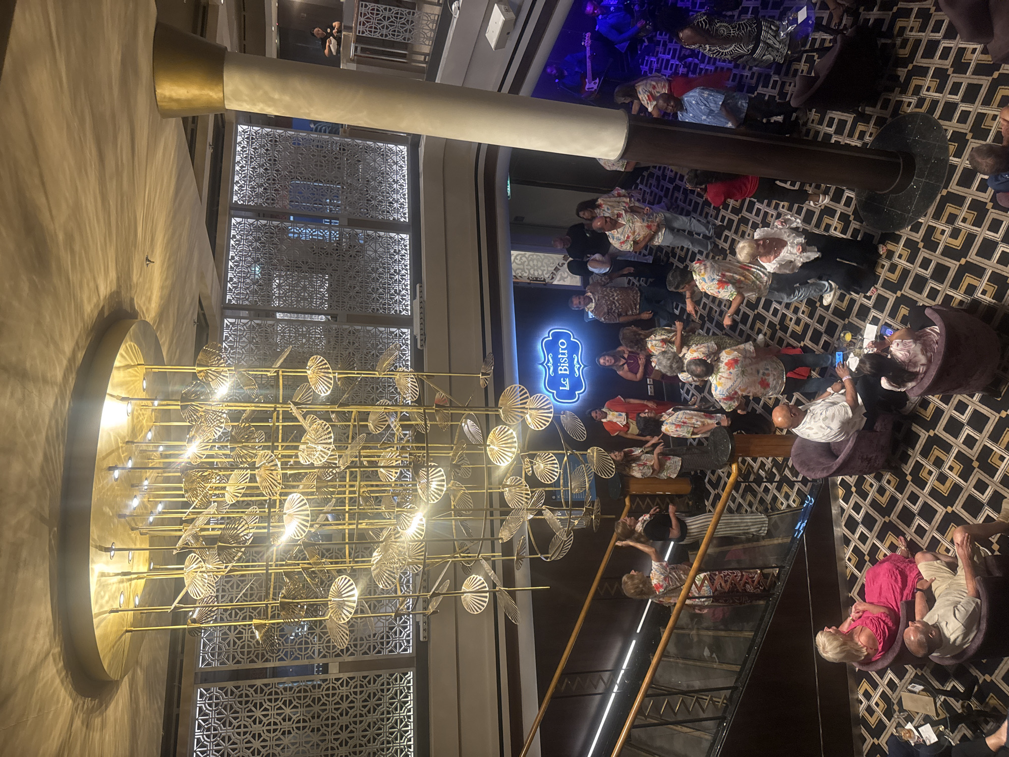 The image depicts an interior scene within what appears to be a cruise ship or a similar entertainment venue. A large, ornate golden chandelier hangs from the ceiling. A group of people are gathered on a lower level, some dancing, others standing and talking. A band is visible on the right, playing instruments. A sign that reads "Le Bistro" is illuminated on the wall. There is a staircase on the left with people standing on it. Several individuals are seated in purple chairs in the foreground. The flooring is patterned with black, gold, and white geometric shapes. The lighting is warm and ambient, creating a festive atmosphere. The location of Motril, Spain is not discernible from the image.