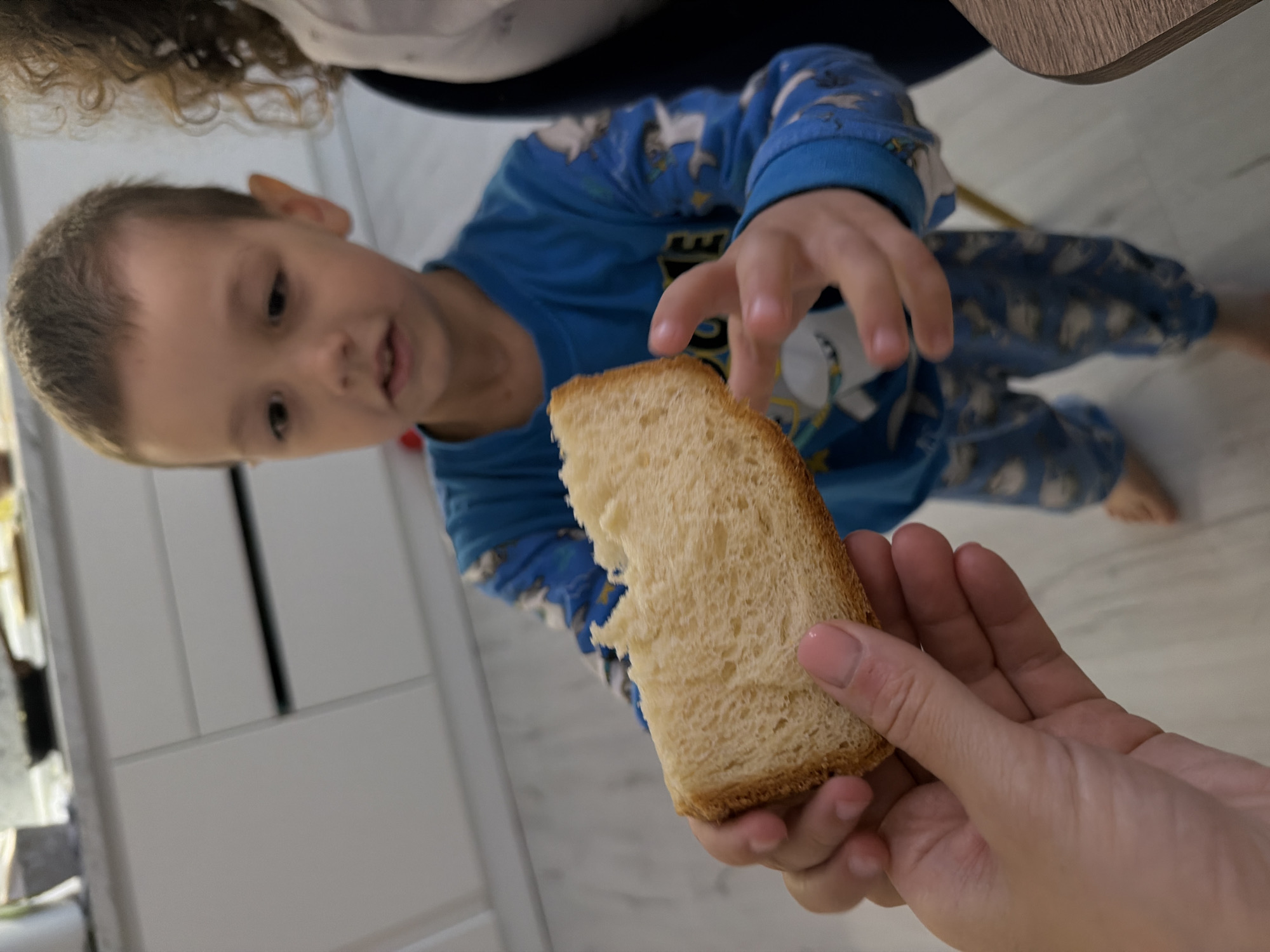 A slightly blurred, medium-shot photo depicts a young child in a blue pajama set with a cartoon print reaching for a piece of bread being held in an adult's hand. The child is looking at the bread with their mouth slightly open. Part of the child's curly hair and shoulder is visible. The adult's hand is in the foreground, holding a golden-brown slice of bread that has a bite taken out of it. The background includes a white cabinet and a tan table. The location and city are not determinable from the image.