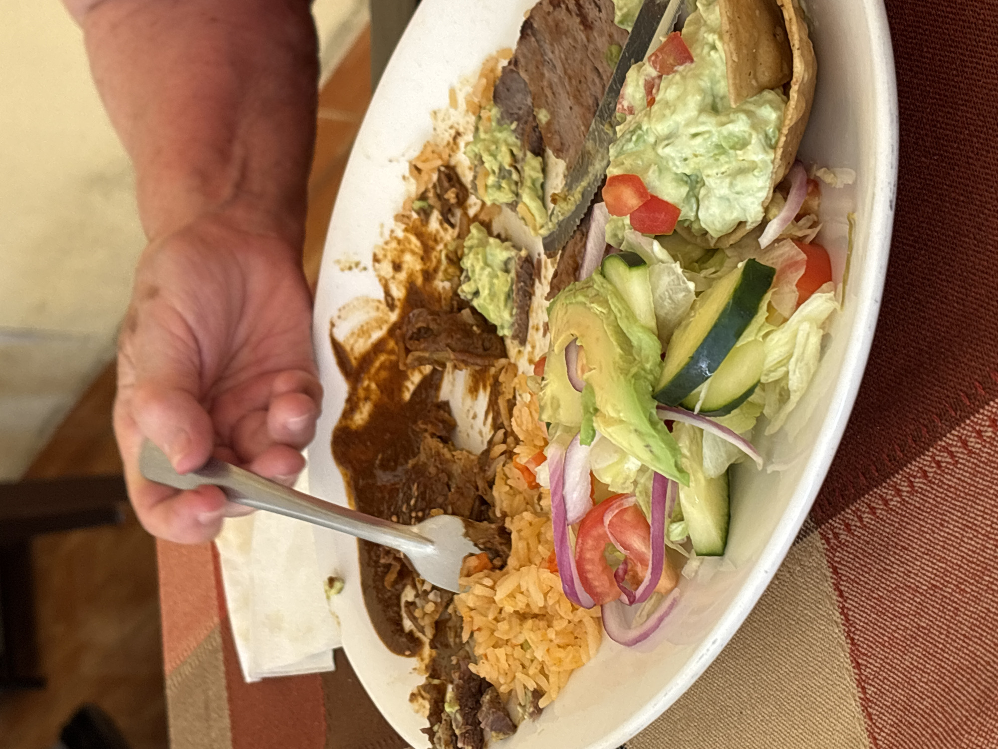 The image presents an overhead shot of a dining scene in San Bernardino Tlaxcalancingo, Mexico, focusing on a plate of food actively being eaten. A person's arm and hand are visible on the left, holding a silver fork that is inserted into a portion of the meal.
The main subject is a white, oval plate filled with a variety of Mexican-style dishes. On the left side of the plate, there are slices of dark meat, possibly carne asada or stewed beef, generously covered in a rich, dark brown sauce, resembling mole. Adjacent to the meat is a serving of reddish-orange Mexican rice. A large scoop of guacamole, flecked with diced red tomatoes, is placed on top of some of the meat and rice, with a piece of a crispy tortilla or tostada shell embedded within it. The right side of the plate holds a fresh salad, consisting of shredded lettuce, slices of cucumber, red onion, tomato, and avocado. A serrated knife is also visible on the plate, resting near the meat and guacamole.
The meal is laid out on a checkered tablecloth featuring patterns in shades of brown, orange, and tan. A white paper napkin is partially visible beneath the person's hand. The background is softly blurred, suggesting an indoor setting, possibly a restaurant or home dining area. The activity captured is a person in the process of enjoying their meal. There is no visible text in the image.