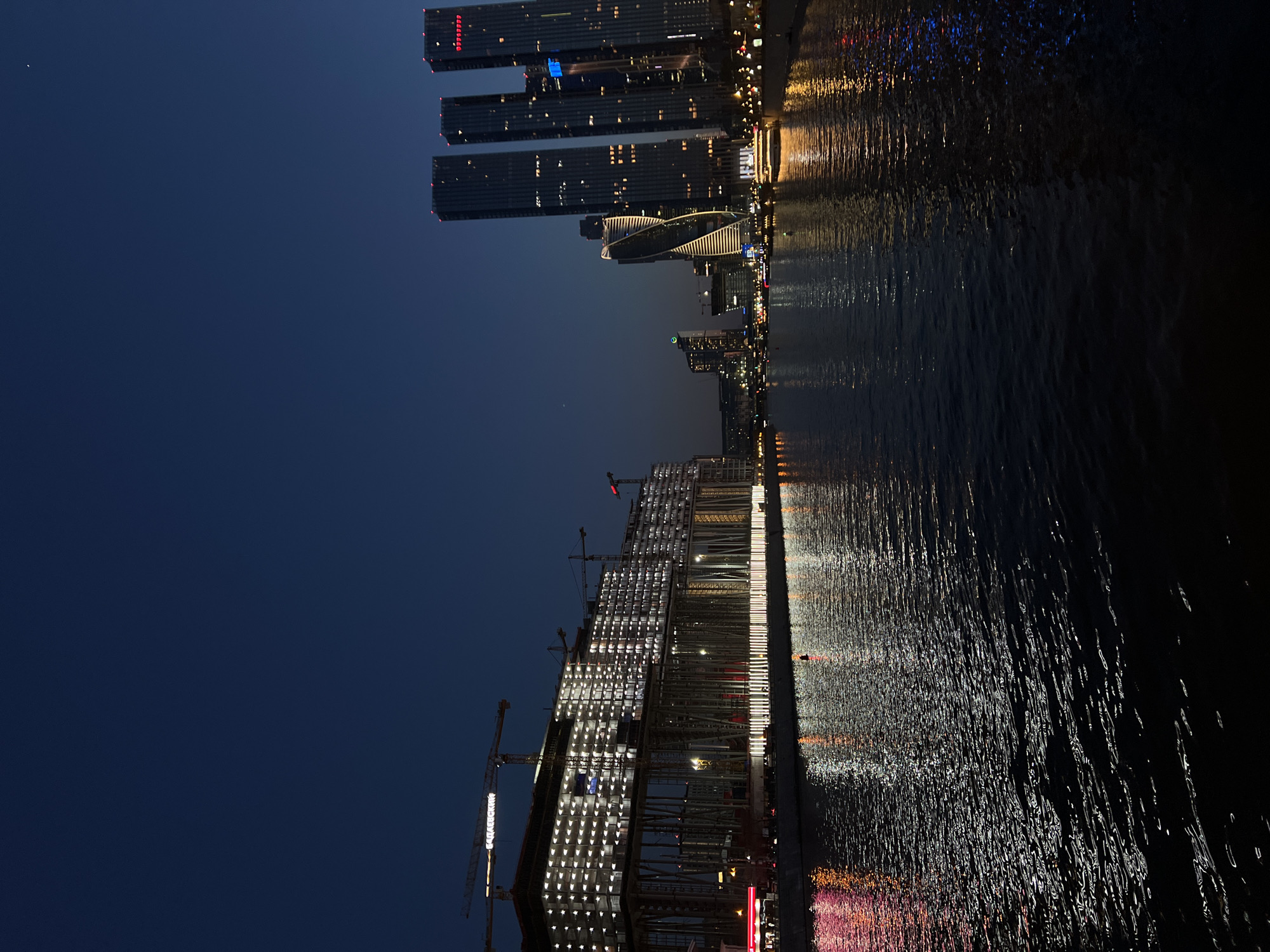 At night, the Moscow International Business Center is captured in this image. The scene showcases a cluster of modern skyscrapers with illuminated windows, their reflections shimmering on the nearby body of water. Under construction, one prominent building features cranes along its lit-up skeletal structure. The sky is dark and clear, providing a backdrop to the city's skyline.