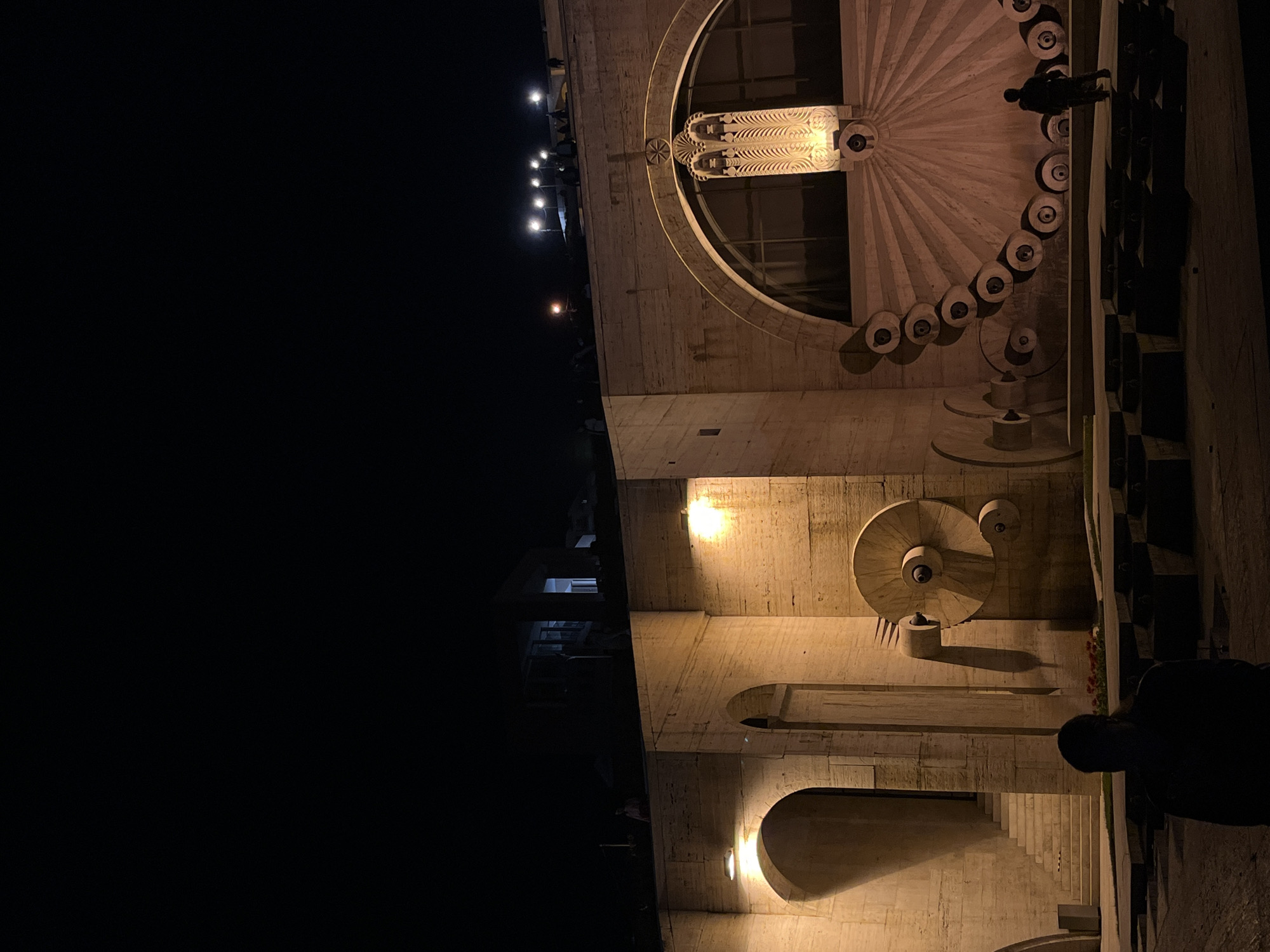 The image captures a monumental, illuminated architectural complex at night, strongly indicative of the Yerevan Cascade in Armenia. The vast majority of the left side of the frame is dark, representing the night sky, while the right side is dominated by the grand structure.
The central subject is a massive, light-colored stone (or concrete) building, likely a multi-tiered staircase or a cascading monument, brilliantly lit by warm artificial lights. Prominent features include a large, ornate circular element towards the top, possibly a window or a decorative facade, adorned with intricate, radiating carvings reminiscent of a sunburst or floral pattern. Below this, a series of smaller, circular, tiered elements descends along a curved path, suggesting water features or decorative fountains, also illuminated.
Further down the structure, arches and rectangular openings create depth, with visible staircases and more wall-mounted lights casting an inviting glow. Another distinct, circular, spiral-shaped decorative or water feature is visible on a lower level.
Several people are present, mostly silhouetted against the illuminated architecture. One prominent figure stands near the upper right, seemingly observing the surroundings. Other indistinct figures are visible further away on various levels of the structure, adding a sense of scale and public engagement.
The setting is clearly nighttime, with the absence of natural light emphasizing the artificial illumination. The weather appears clear, with no atmospheric elements obstructing the view. The overall atmosphere is grand and serene, showcasing the architectural beauty in a dramatic light. No discernible text is visible on the structure.