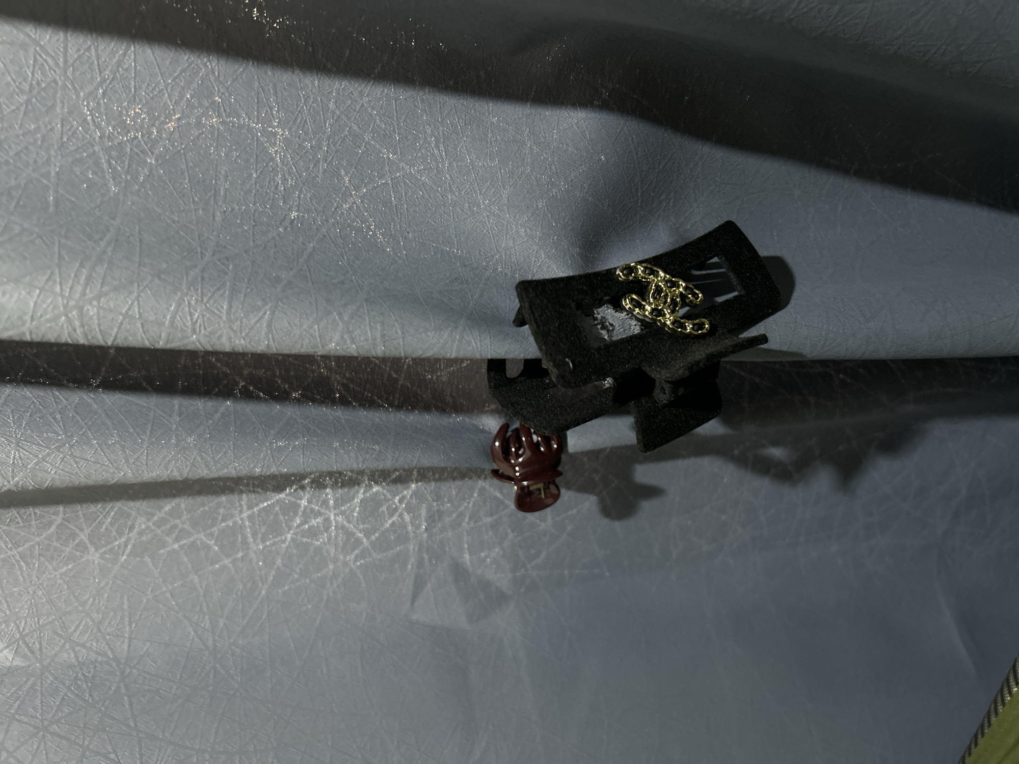 A dark brown hair clip is attached to a dark grey textured curtain. The hair clip is holding a small black object, possibly a pouch or case, which has a gold chain attached to it. The chain is looped and draped over the edge of the black object. The curtain is lit from the right, casting a shadow to the left. The overall impression is of a dimly lit indoor scene, potentially a bedroom or dressing area. The location in Sukawati, Indonesia is not discernible from the visual information provided.