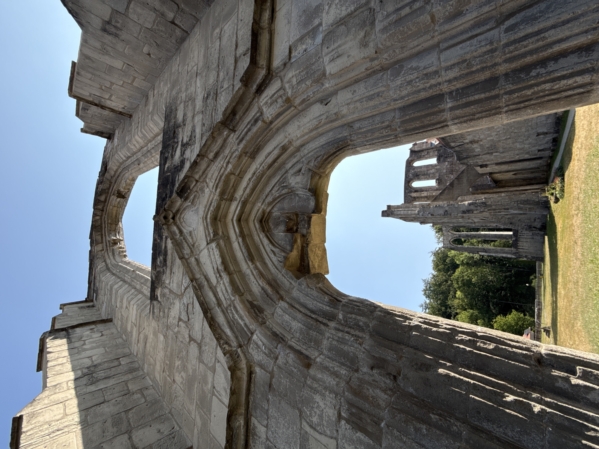 The image shows the ruins of the Walkenried Abbey in Walkenried, Germany. The perspective is from below, looking up at the stone architecture against a clear blue sky. The foreground is dominated by a large, arched stone structure, with visible masonry and decorative carving. Light and shadow play across the stone, highlighting its textures and the architectural details.
Beyond this foreground archway, the ruins of the abbey extend into the background. We can see more stone walls, arched windows, and remnants of the building's former grandeur. Green trees and foliage surround the ruins, and a grassy field is visible in the middle ground, with a hint of a pathway and some landscaping. There are no people clearly visible in the image. The scene conveys a sense of historical preservation and architectural decay in an outdoor setting.