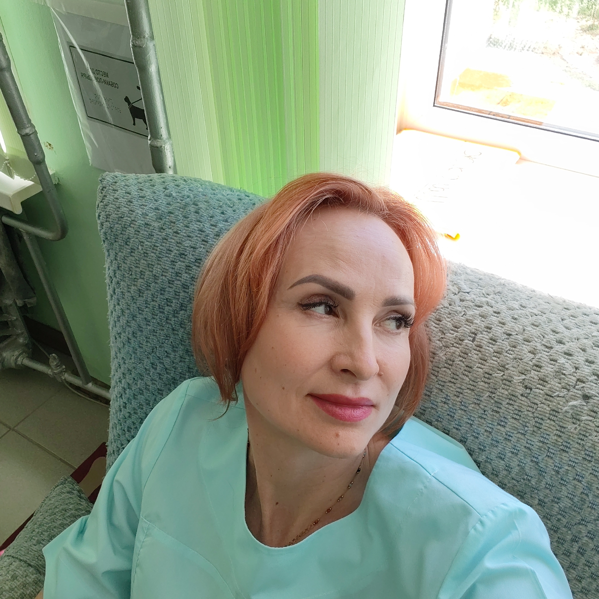 A woman with short reddish-orange hair is seated in what appears to be an upholstered chair. She is wearing a light blue uniform-style top. The background includes light green vertical blinds, a window with bright light, and a metal frame structure partially visible to the left. There's also a sign attached to the structure with an image of a dog on it. The location is in Chaikovsky, Russia, based on the text written on the sign.