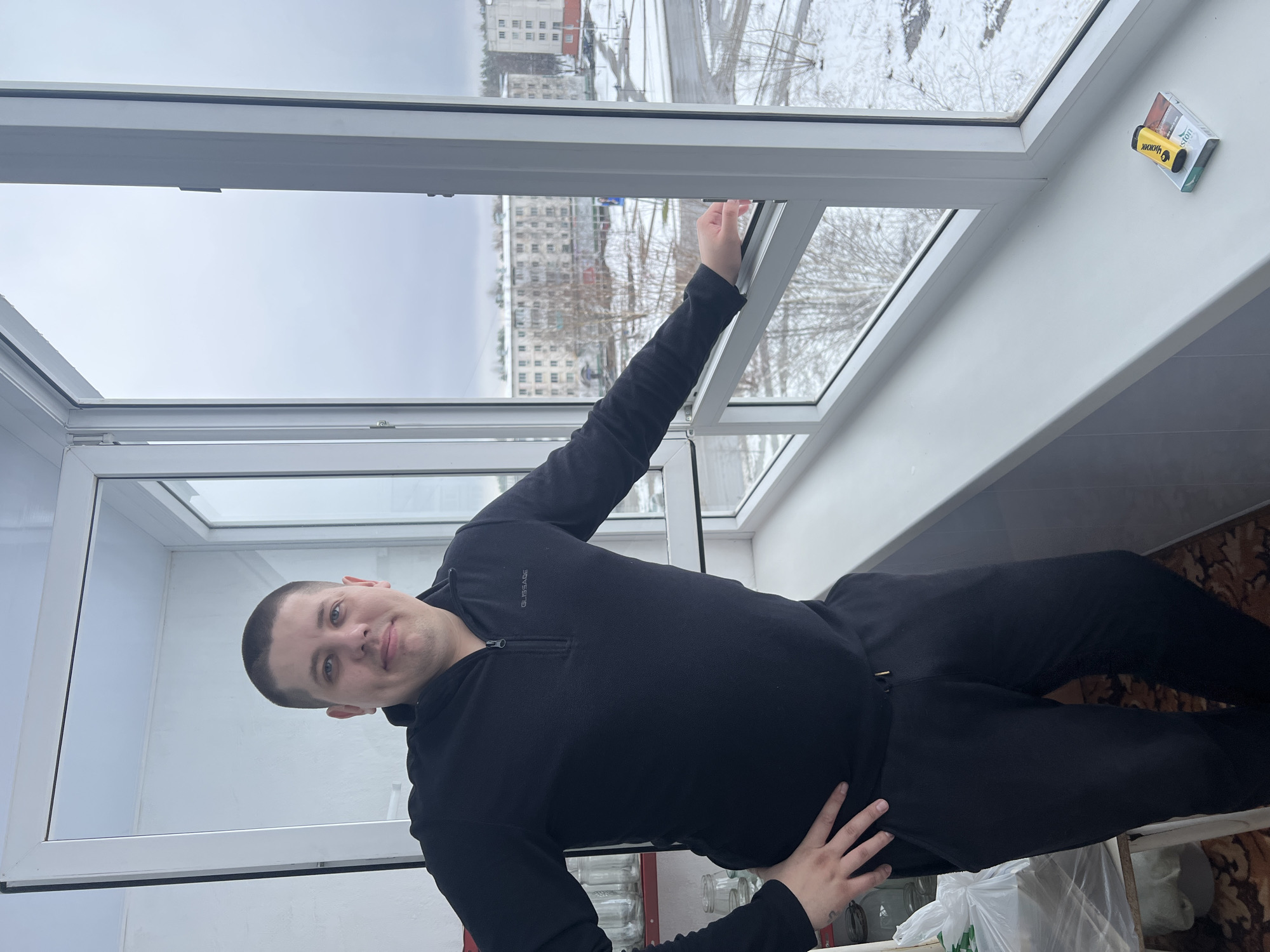 A man in a black long-sleeved shirt and black pants is standing in front of a white window, looking at the camera. His left hand is on his hip, and his right arm is extended upwards towards the window. Outside the window, a city scene is visible with buildings and bare trees, likely taken during winter given the presence of snow. Based on the objects in the room, including the jars and bottles, it appears he may be in a kitchen or a storage room. The location is Chaikovsky, Russia, based on the "Чайковский" branded item stuck to the window.