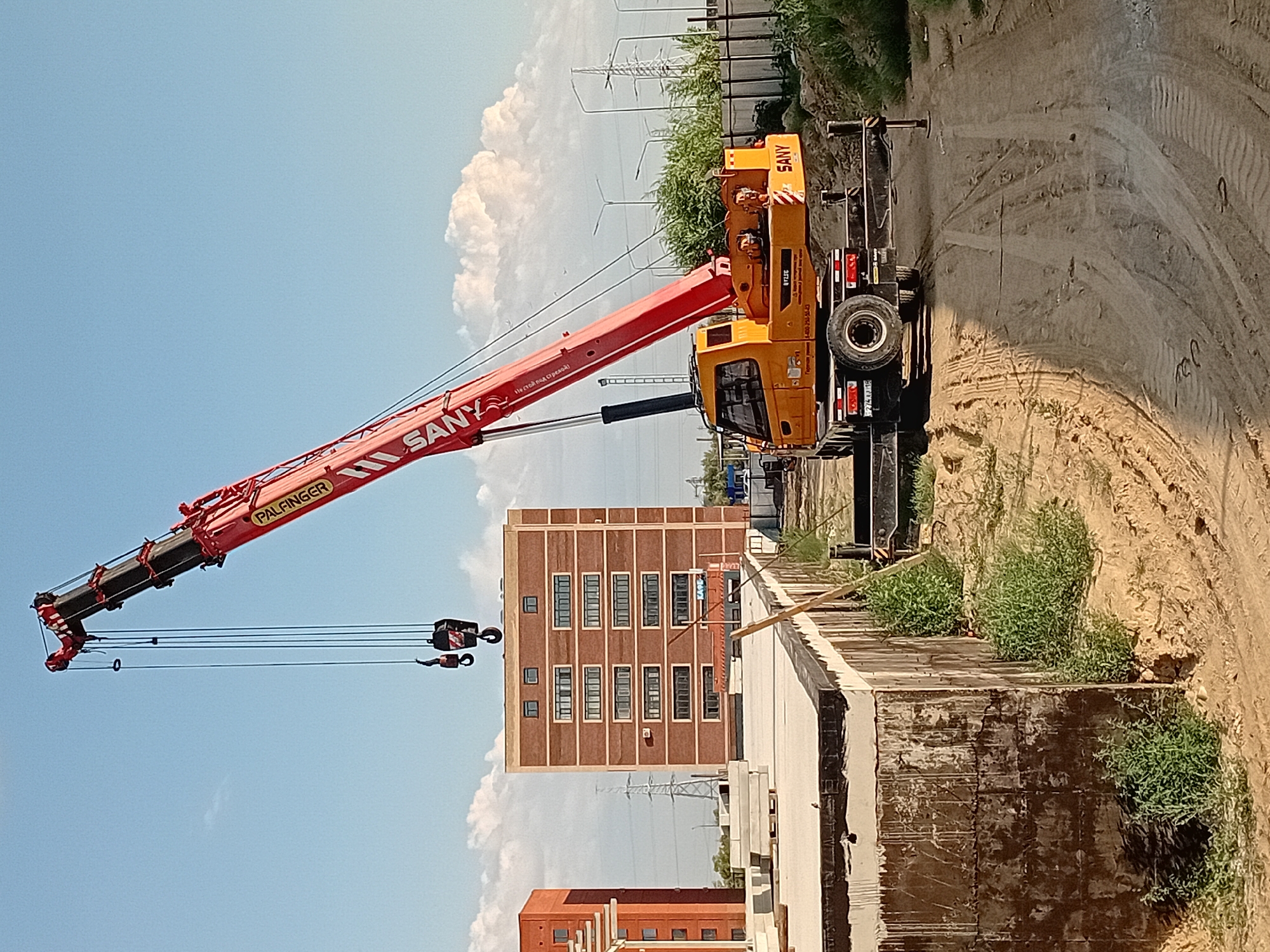 A construction site in Semey, Kazakhstan features a large, red mobile crane with "PALFINGER" and "SANY" branding, its boom extended upwards and to the right. The crane is positioned on a dirt road, with its yellow cab visible. In the background, two multi-story brick buildings stand under a partly cloudy blue sky. One building is predominantly red-brick with white trim and numerous windows. The other, to its left, is also red-brick. Various construction materials, including stacked concrete slabs, are visible on the ground. Green vegetation, including bushes and grass, grows around the site. Electrical transmission towers and streetlights are also present in the distance, indicating an urban or semi-urban setting. The overall scene depicts an active construction zone.