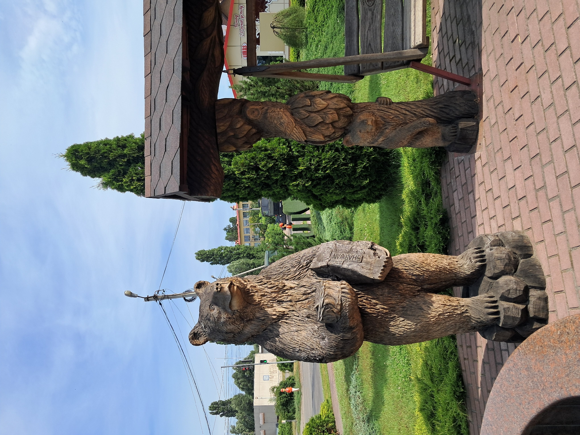 The image shows a wooden sculpture of a bear holding a book in its paws, standing on a brick pathway in Kremenchuk, Ukraine. The bear sculpture is detailed, with carved fur and claws. Behind the bear, to the right, is a wooden structure with a brown, shingled roof and a bench swing. The structure is supported by a wooden pillar that is also carved with figures, including an owl and another bear cub. In the background, there are green trees, a grassy area, and buildings. A street light is visible above and behind the bear. The sky is blue with some scattered clouds.