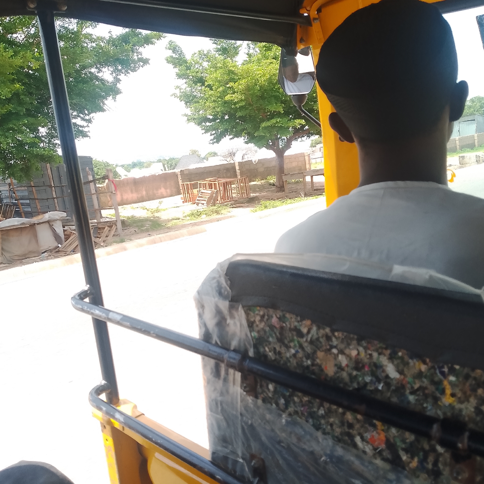 The image shows the interior of a yellow auto-rickshaw, likely in Jalingo, Nigeria. The view is from the back seat, looking forward through the open sides of the vehicle. A person wearing a white shirt and a black cap is seated in the front, facing away from the camera. The background reveals a street scene with trees, brick walls, stacks of building materials, and what appear to be wooden structures. Sunlight illuminates the scene, suggesting it is daytime. The seat in the foreground is covered with a transparent plastic sheet, revealing a colorful, textured material underneath, possibly a seat cushion or padding filled with debris. The overall environment suggests an urban or semi-urban setting.