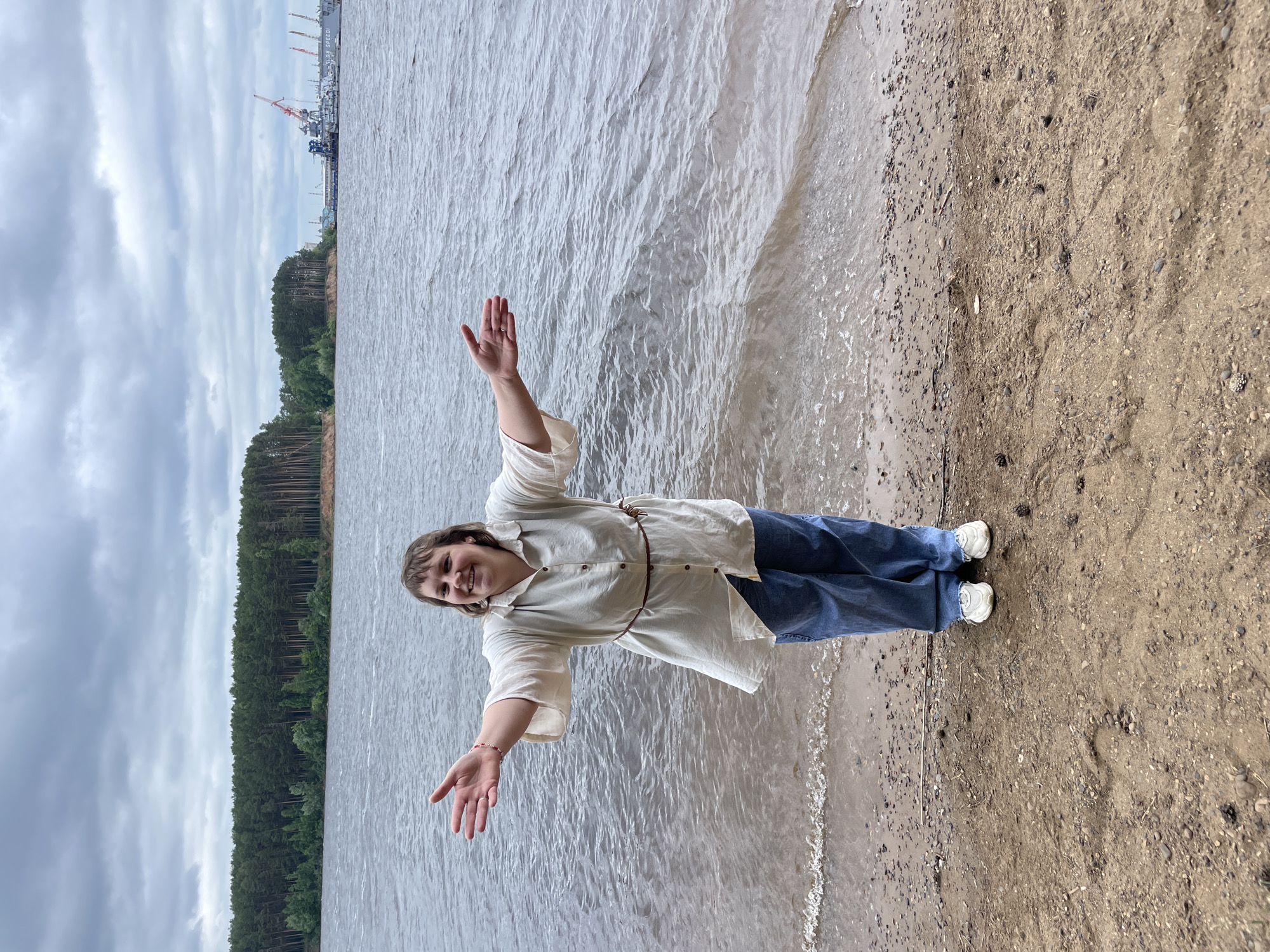 A person stands on a sandy shore by a large body of water, with arms outstretched. They are wearing a light-colored, long-sleeved tunic over blue denim jeans and white sneakers. A thin brown belt is cinched around the waist of the tunic. The person has short, brown hair and is smiling. Behind them, the water is choppy under an overcast sky. In the distance, across the water, a dense line of pine trees forms the shoreline. Further to the right, industrial structures including cranes and docked vessels are visible. The location is identified as the Kama River in Chaikovsky, Russia.