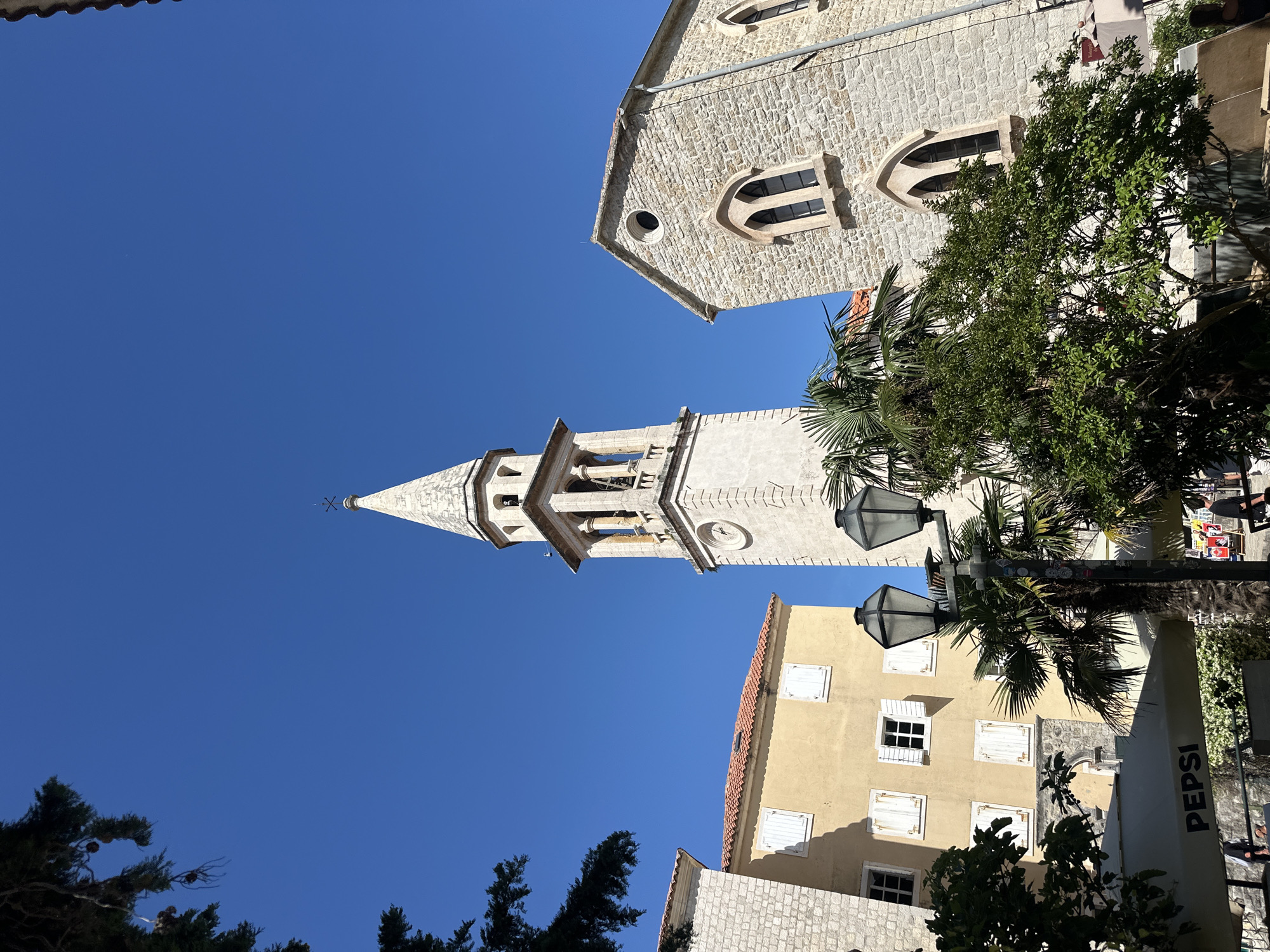 The image shows a low-angle view of buildings and architectural elements in Budva, Montenegro. The central element is a tall, white, stone bell tower with a pointed steeple, topped with a cross. To the right of the tower is a building constructed of light-colored stones with distinctive arched windows. Below and slightly to the right is a yellow building with white-shuttered windows. Palm trees and other greenery are interspersed between the buildings. A black lamppost with two lanterns is partially visible in front of the yellow building. The sky is a clear, bright blue. Part of a "PEPSI" sign is visible on the right. The composition suggests a narrow street or square in the old town of Budva.
