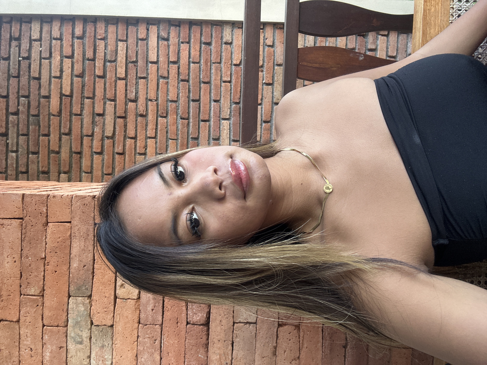 A woman is lying down outside in Ubud District, Indonesia. The woman is lying on a wooden chair with a lattice seat. She is wearing a black top and a gold necklace. Her hair is black and blonde. The woman is lying next to a brick wall. A similar brick wall is also behind the wooden chair.