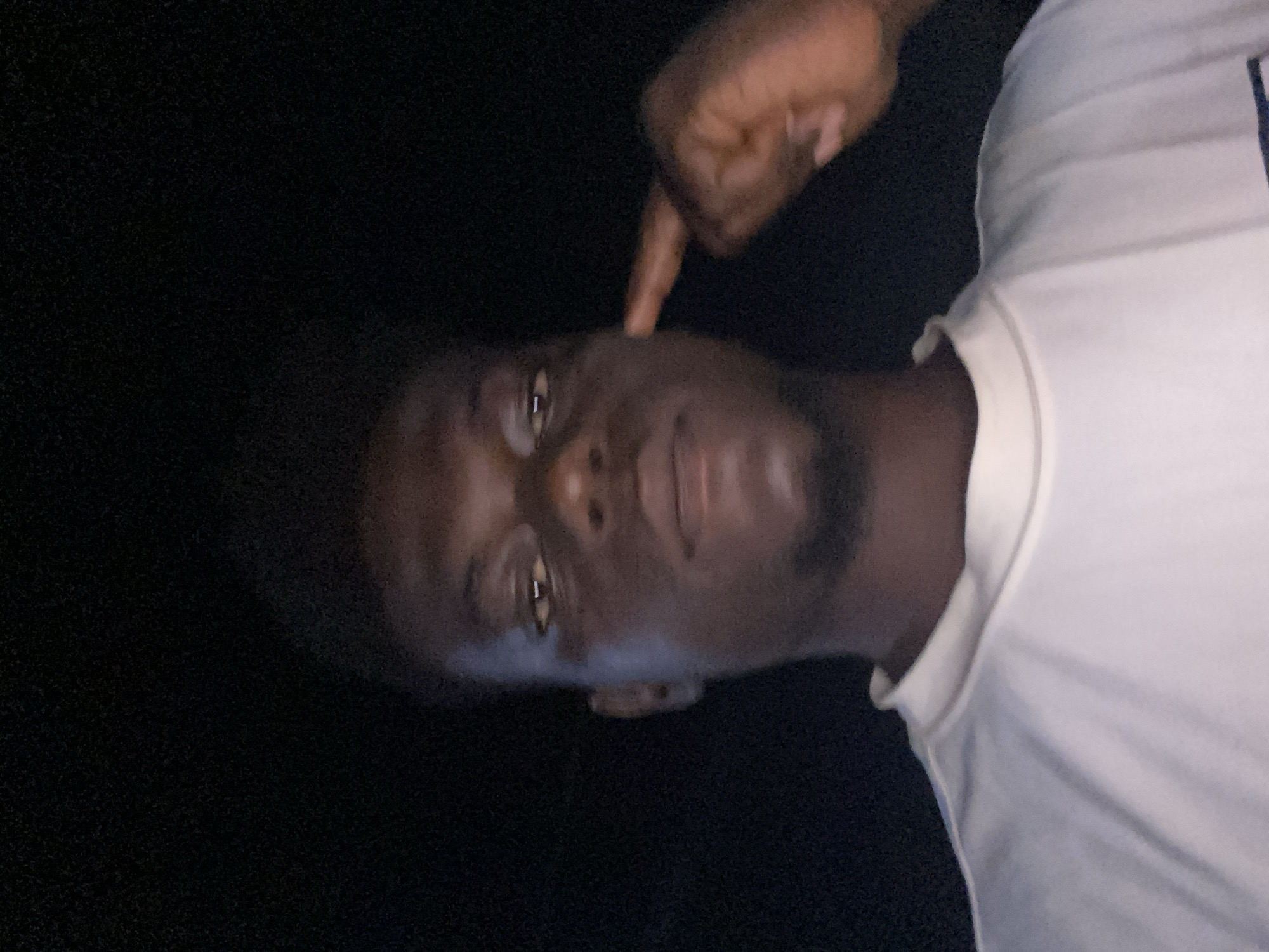 A person with dark skin and short dark hair is pointing to their cheek with their right index finger. The person is wearing a white shirt. The background is dark, suggesting a nighttime or dimly lit environment. The location is Unknown, Unknown.