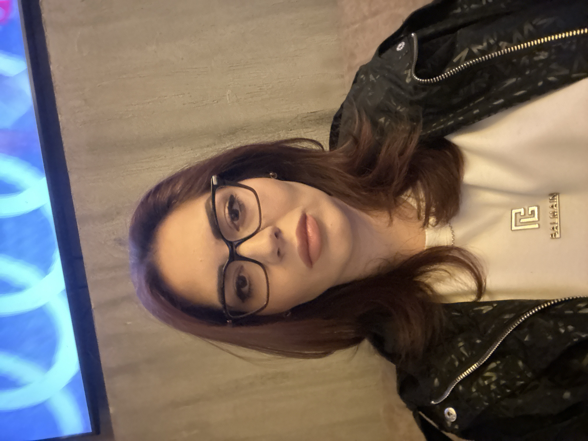 A woman with reddish-brown hair and glasses is posing for a portrait indoors. She is wearing a white t-shirt with a silver logo and a black jacket. To the left of the frame, a television screen displays abstract blue and white patterns. The background consists of a textured grey wall. The location is indeterminate and cannot be definitively placed in Sofia, Bulgaria based solely on the image.