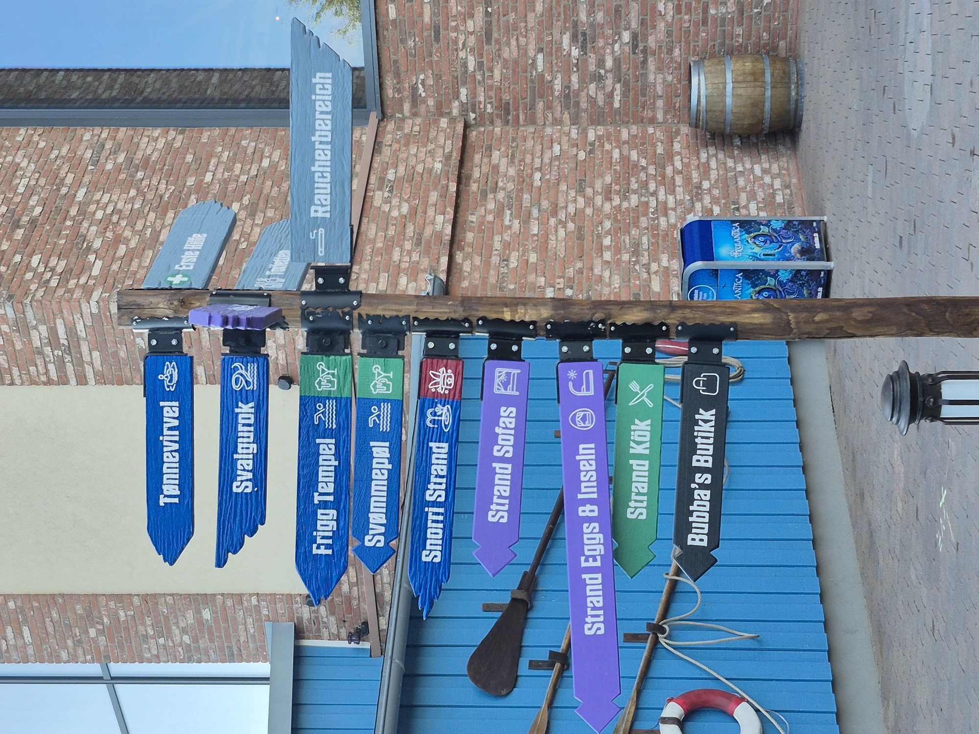 In Rust, Germany, an outdoor signpost with multiple directional arrows is visible. The signpost is made of wood and stands on a paved area. The arrows are painted in various colors: blue, purple, green, and black. Some arrows have text and icons. Examples of text include "Tønnevirvel," "Svalgurok," "Frigg Tempel," "Svømme pøl," "Snorri Strand," "Strand Sofas," "Strand Eggs & Inseln," "Strand Kök," and "Bubba's Butikk." Some arrows also point to "Erste Hilfe" and "Raucherbereich." To the left of the signpost is a blue building with wooden planking. An oar is mounted on the blue wall. A red and white life preserver is also visible. To the right of the signpost, there is a blue trash receptacle with images of animated fish, and a wooden barrel. The background consists of a brick wall and a window. The sky is visible at the top of the image.