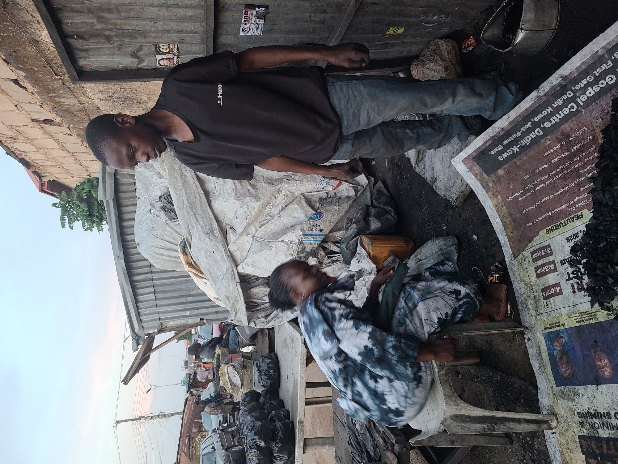 This media file captures an outdoor scene, likely a street or market setting, in Jos, Nigeria, during what appears to be dusk or early evening, indicated by the soft, light blue sky.
In the foreground, a man stands wearing a dark t-shirt with "Hero" visible on it and blue jeans. He is looking downwards, with his hands appearing somewhat soiled. Beside him, on the ground, are large white sacks, one of which has "POLYETHYLENE 85 kg M" and "بروج" (Borouj) printed on it. A woman is seated on a low stool or chair to the right, wearing a patterned blue and white garment. She is also looking down, possibly at something in her lap or on the ground.
The ground is rough and uneven, with dirt, stones, and black bags, possibly filled with refuse or charcoal, scattered around. To the right of the woman, a large sheet of paper, possibly a newspaper or poster, is laid out, covered with a pile of dark, coarse material, likely charcoal. Behind the individuals, there are corrugated metal sheets forming part of a makeshift structure, and further in the background, other people and structures can be vaguely seen, suggesting a bustling environment. A small, yellow container is also visible near the woman.
Visible text on the large paper includes details for an event: "Gospel Centre, Dadin-Kowa, First Gate, Dadin Kowa, Jos-Plateau State." It mentions "FEAUTURING" various "Topics On: ways to Academic Excellence," "Kingdom Wealth and Financial minion," "marriage and sexual purity," and "How to know your partner before committing." Event details include "SATURDAY MORNING - Football Match." and "SUNDAY - Grand Finale," with specific times like "3:30pm," "4:00?14," and "6:30am," and a date "2025." Other small posters on the corrugated metal show "OBELE Birthday" and "ABARI."