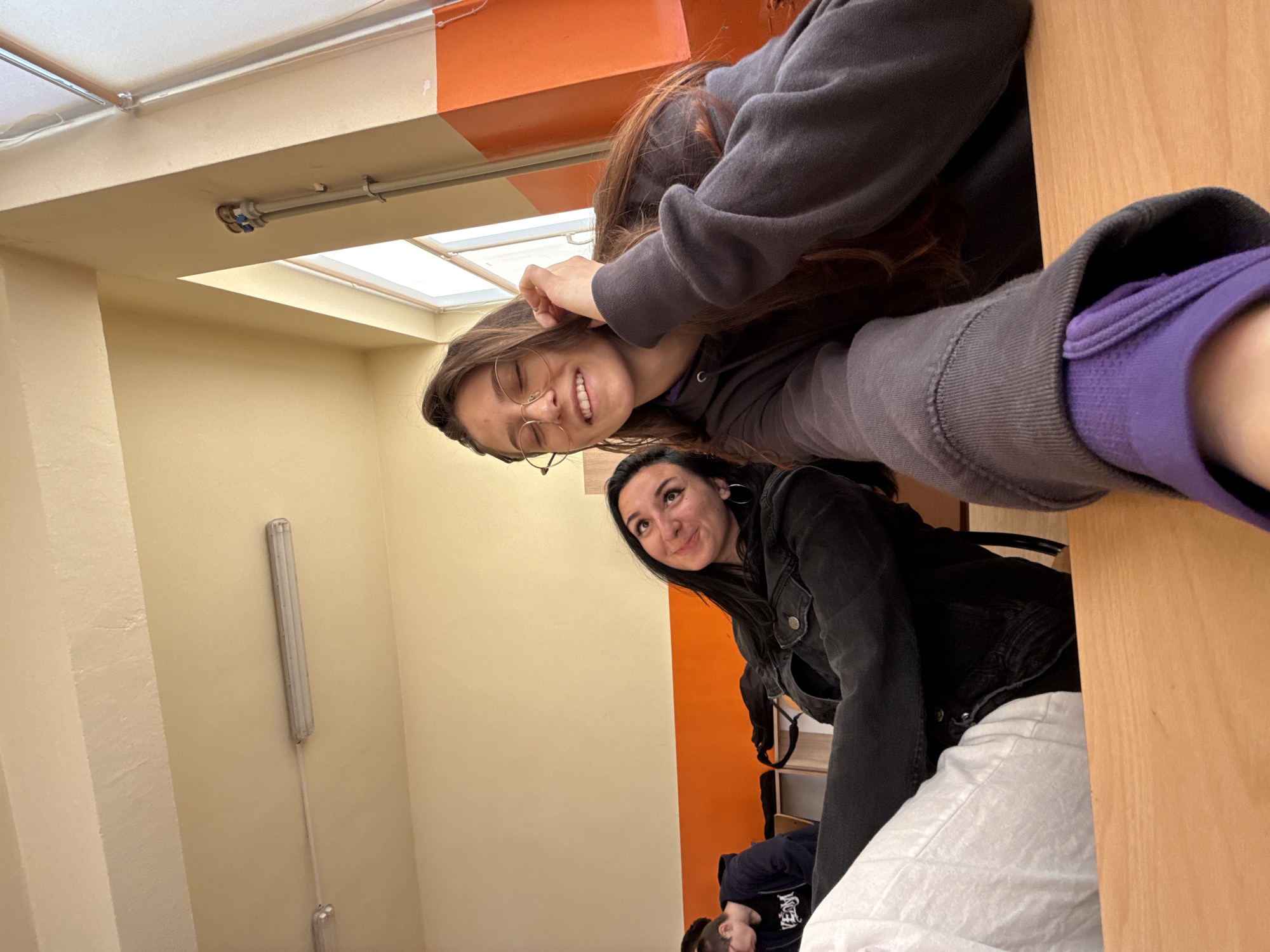 Two young women are posing for a picture in what appears to be an indoor setting with orange and beige walls and a drop ceiling. One woman, wearing glasses and a gray hoodie with purple lining, is smiling and looking at the camera. Another woman, wearing a black jacket and white pants, is looking slightly upward. A light fixture is visible on the wall. A third person is partially visible in the background. The location is likely within the city of Sofia, Bulgaria, based on the environment.