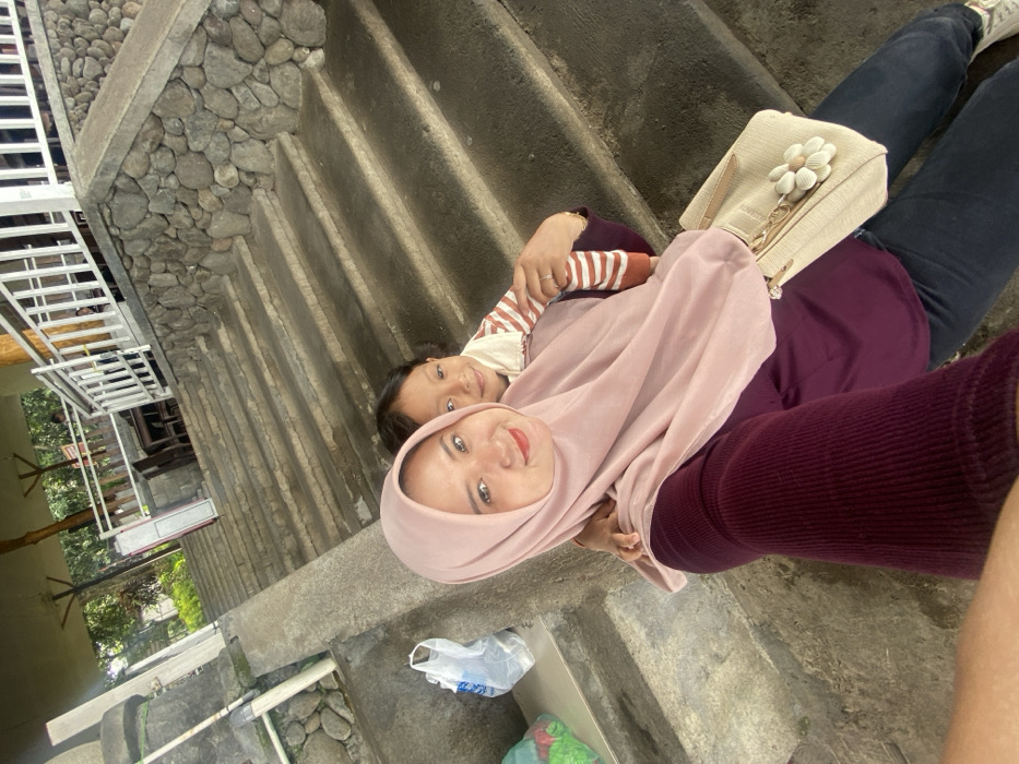 A woman wearing a light pink hijab and burgundy sweater sits on stone steps with a young child wearing a striped shirt. The woman is smiling and holding the child close. A beige purse with a flower embellishment sits beside them. The steps are wet and there is trash scattered nearby. A metal railing and greenery are visible in the background.