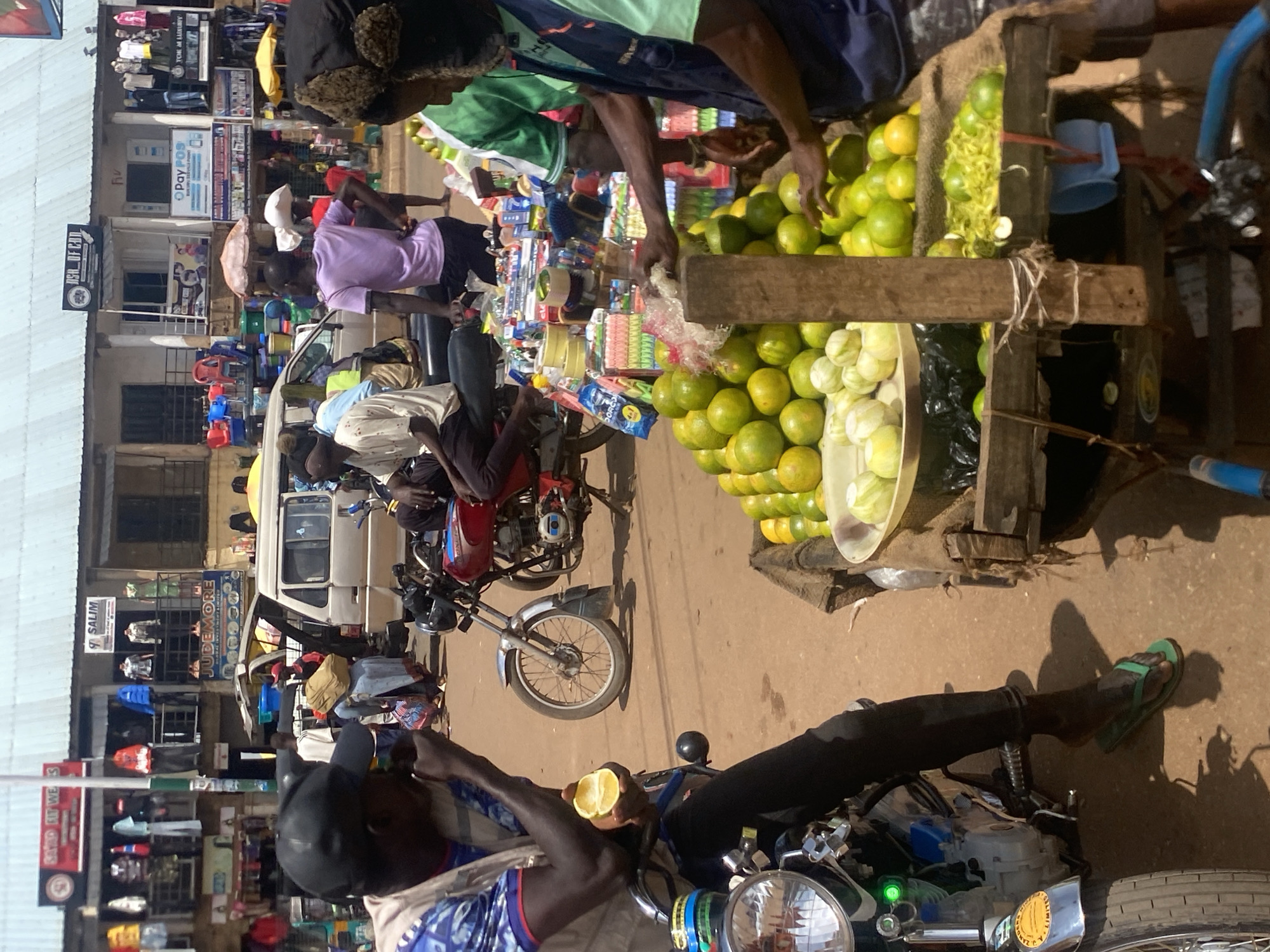 In the city of Bukuru, Nigeria, a street scene unfolds with a bustling market environment. In the foreground, a man wearing a black baseball cap and a blue and white patterned shirt is seated on a motorcycle, holding a halved lemon in his right hand, appearing to be eating or preparing to eat it. His black pants and green flip-flops are visible. To his right, a produce stand is laden with a variety of fruits, predominantly oranges and other citrus varieties, in shades of green and yellow. Chopped green produce is also visible.
Behind the man and the motorcycle, a street market is in full swing. Several people are milling about. One man in a purple t-shirt and black pants is walking with a bundle on his head. A red motorcycle is parked nearby. In the background, shops line the street, displaying clothing on racks and mannequins. Various signs are visible, including "SAYID FIT WEAR" and "SALIM." A white van is also present in the market. The ground is dusty, indicative of the dry, outdoor environment. The overall scene depicts a typical day in a Nigerian market, filled with commerce and daily life activities.