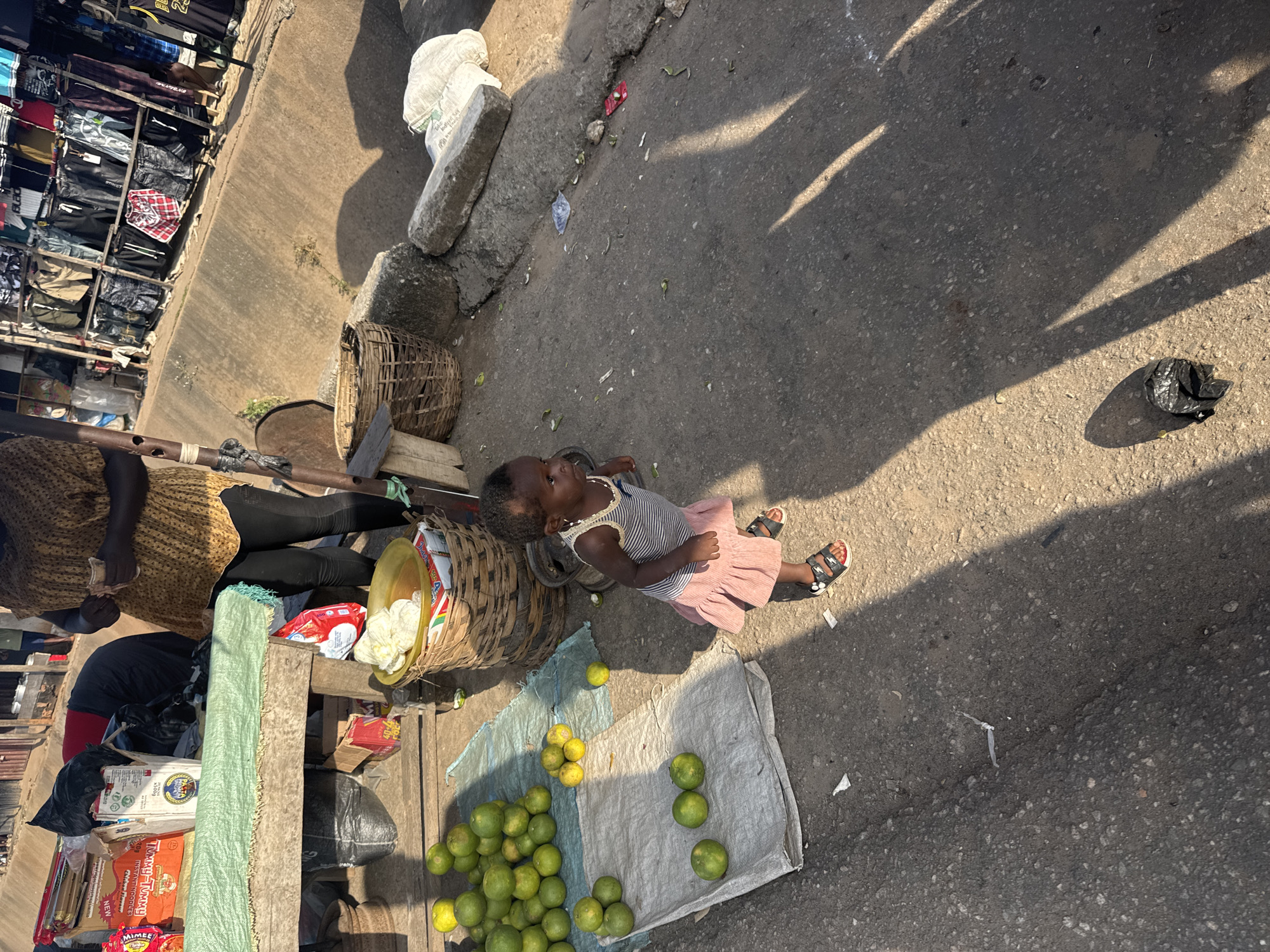 This image depicts an outdoor market scene, likely in Onitsha, Nigeria. The primary subjects are a young child and an adult, likely a vendor. The child, wearing a striped top and a pink skirt, is looking upwards, possibly at the sun or someone off-camera, with a neutral expression. The adult, dressed in a patterned top and dark leggings, is partially visible and appears to be handling money or goods.
The setting is a bustling marketplace with various items for sale. To the left, shelves are packed with clothing, primarily t-shirts and pants. In the foreground, there's a stall with goods, including packaged food items like "TOMMY-MAMY" instant noodles and "MIMEE" crackers. Bags of produce, possibly oranges, are displayed on a cloth on the ground. Woven baskets are also present, one containing some white items.
The ground is paved with asphalt, suggesting a street market. Sunlight casts long shadows across the scene, indicating it's likely daytime, possibly late morning or afternoon. The presence of loose produce and some litter on the ground contributes to the gritty, authentic atmosphere of a busy market. The overall mood is one of everyday commerce and activity. No specific event or urgent activity is evident, but rather a snapshot of daily life in a Nigerian market.