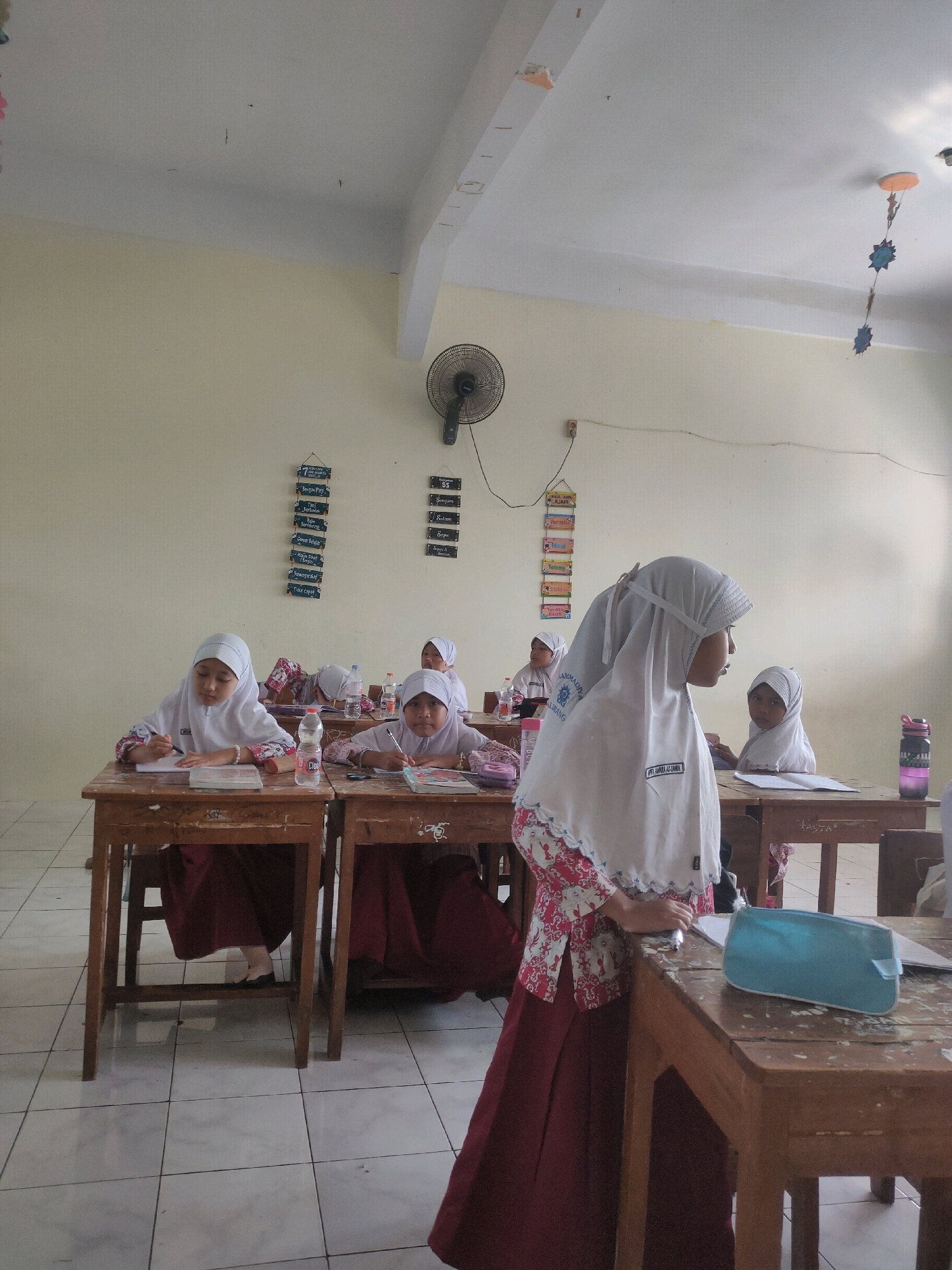 The image depicts a classroom scene at a school in Payaman, Indonesia. Several young female students, dressed in white hijabs and maroon skirts, are seated at wooden desks, engaged in learning activities. One student in the foreground is standing and looking to her right, while others are seated and writing or reading. The classroom walls are painted a light yellow, and there are educational posters and decorations on the walls. A ceiling fan is visible on the wall. The floor is tiled. The scene appears to be during a school day, with students focused on their studies.