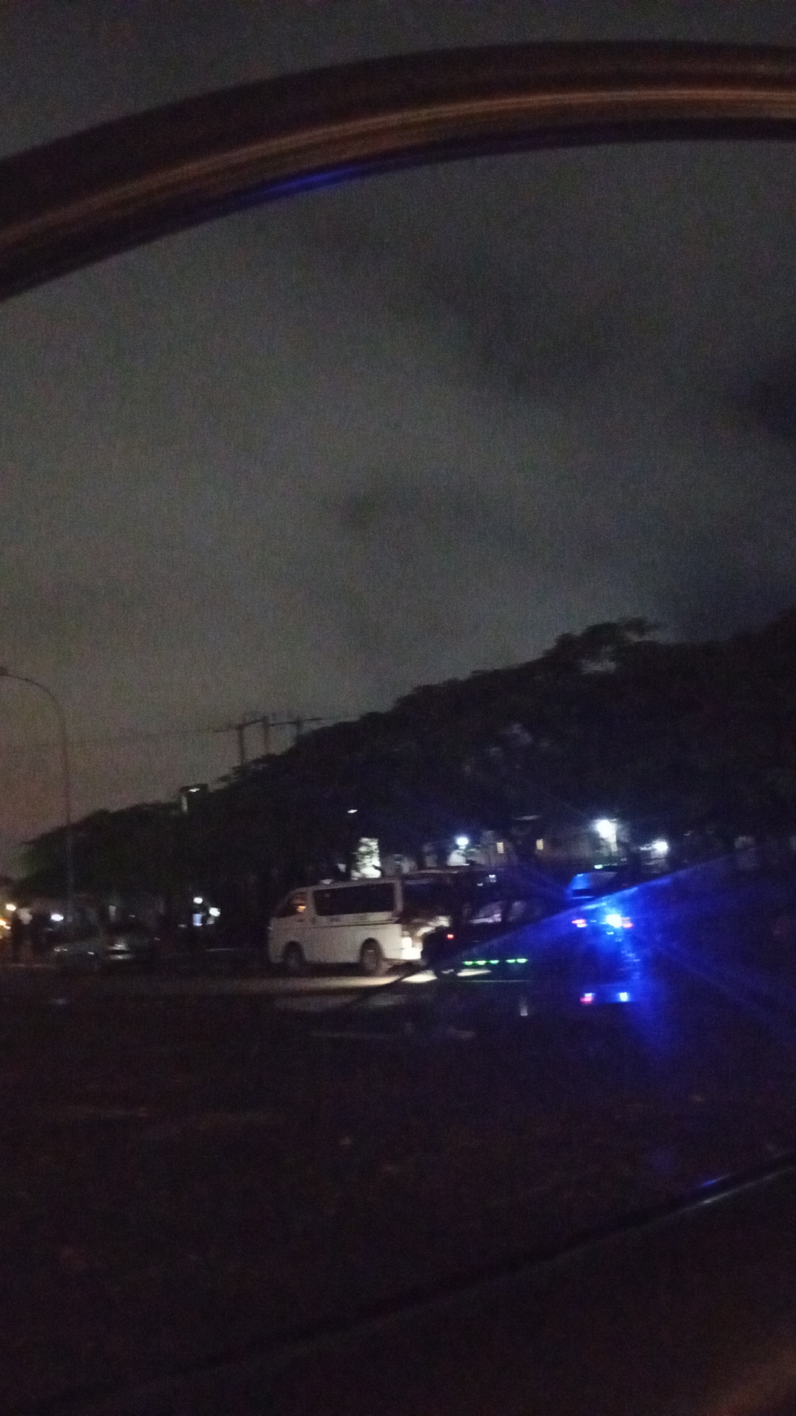 The image depicts a nighttime scene in Aso, Nigeria, viewed from inside a vehicle. In the foreground, a dark, reflective surface, likely a window or a mirror, partially obscures the view. The middle ground shows a street with several vehicles. A white van is parked on the right side of the frame, illuminated by artificial light. To its right, a dark-colored vehicle with bright blue lights is visible, suggesting it might be an emergency vehicle or has aftermarket lighting. In the background, a dark, treeline rises, and behind that, a faint silhouette of buildings or structures is discernible against a cloudy, dark sky. The overall atmosphere is dim, with artificial lights from the vehicles creating bright spots in the darkness.