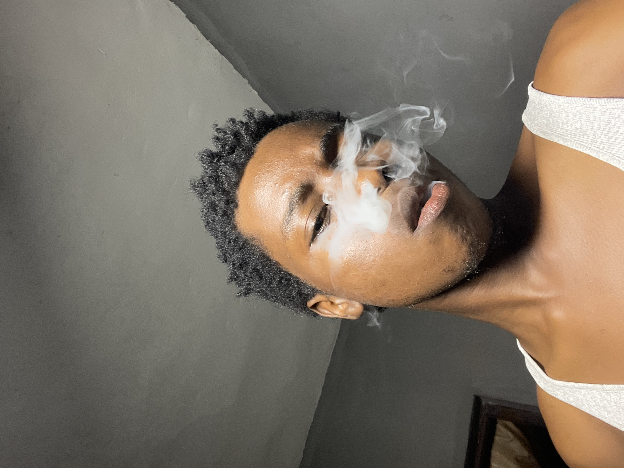 A young Black individual is shown from the chest up, facing slightly to the right with their eyes looking towards the viewer. They have short, curly dark hair and are exhaling a cloud of white smoke, which obscures part of their face. The individual is wearing a white tank top. The background appears to be an interior space with gray walls and a ceiling. The lighting is dim, casting shadows on the individual's face and the surrounding environment. The image is taken from a low angle, looking up at the individual. There are no clear indications of the specific location within Nkpor, Nigeria, beyond it being an indoor setting.
