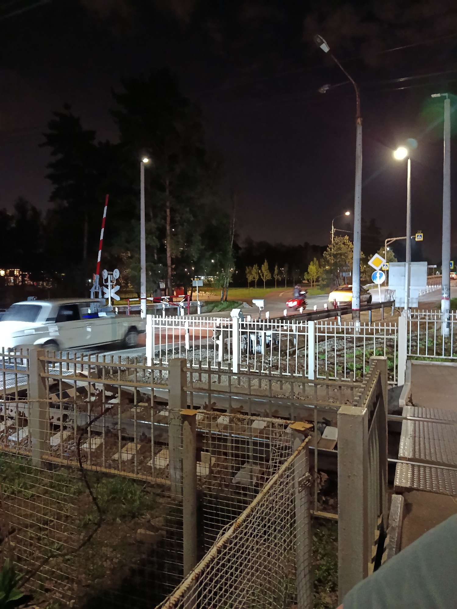 The image depicts a railway crossing at night. A white car is crossing the tracks while a red motorcycle waits on the other side. The crossing gate is down, with red and white stripes visible. There are streetlights illuminating the area, and road signs are present. The scene is framed by metal fences and a view down to the tracks themselves. The location is Barvikha, Russia.