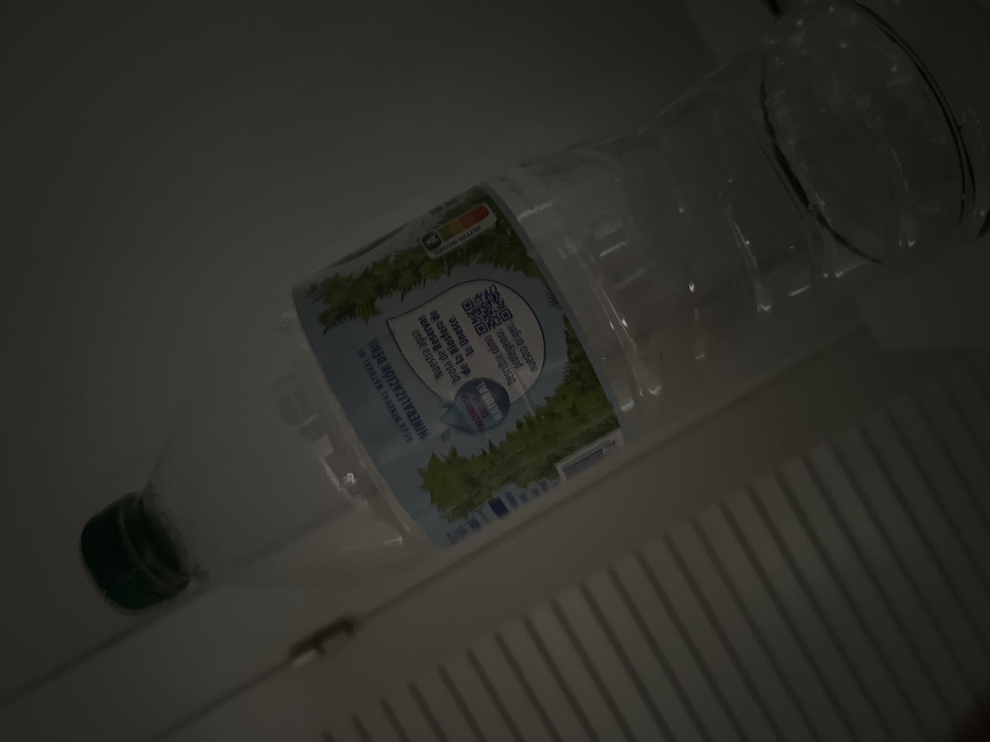 The image shows a plastic water bottle with a green cap, lying on its side. The bottle has a label with text and graphics. The label features an illustration of trees and a landscape, along with nutritional information labeled "NUTRI-SCORE". There is also a QR code. Part of the text on the label is visible and reads "AGUA MINERAL NATURAL DE MINERALIZACIÓN DÉBIL" and "Nuestra agua RECORECTA brota de Reservas NATURAL de la Biosfera de La Unesco. Descubre cómo protegemos nuestro origen:". The image is dark, with the bottle being the primary illuminated subject. The background appears to be a white surface with vertical lines, possibly a refrigerator or a similar appliance. The lighting suggests it might be indoors. The overall scene is a close-up of the water bottle, angled from the bottom left to the top right of the frame.