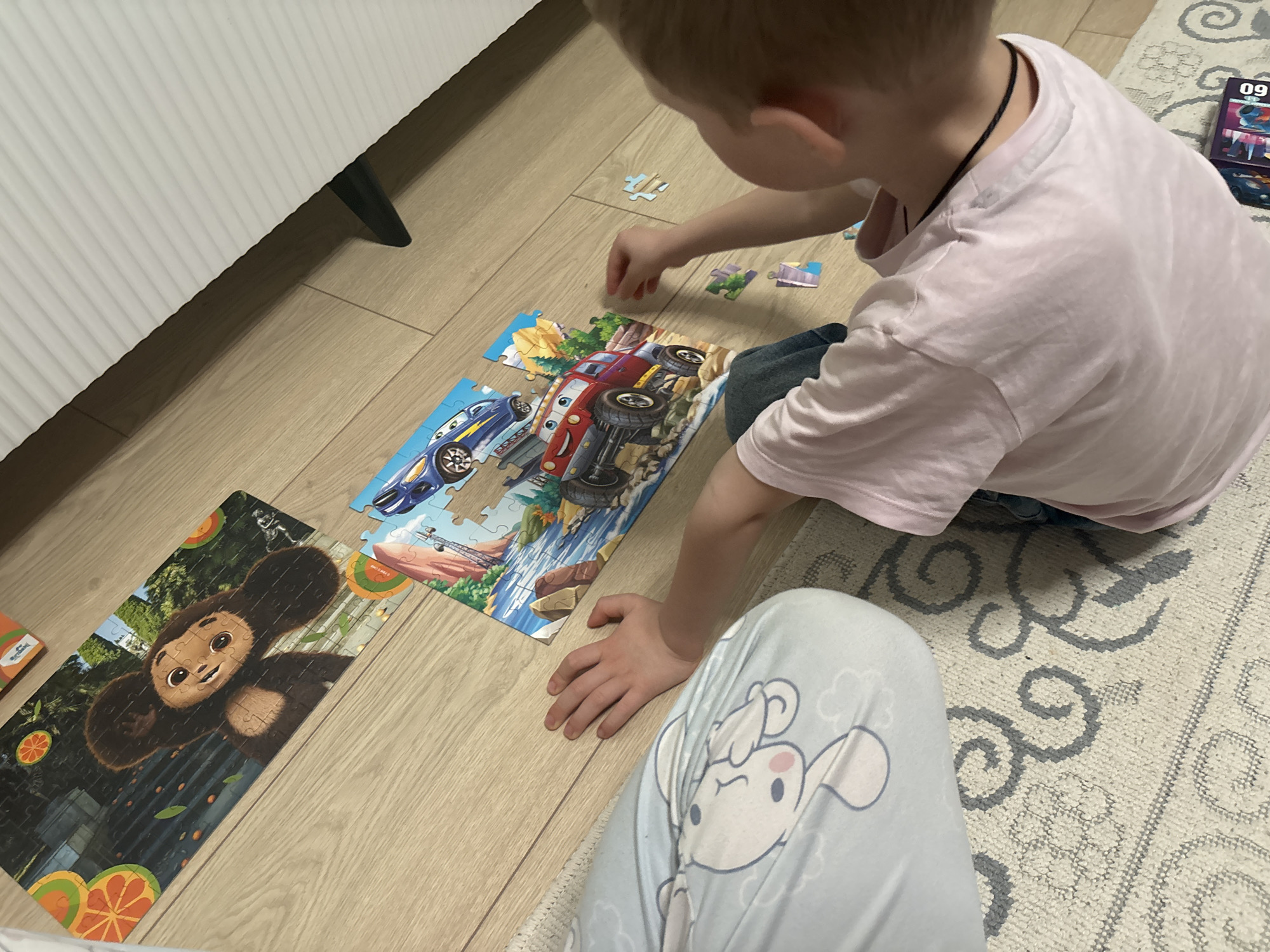 A young child is sitting on the floor, assembling two separate jigsaw puzzles. One puzzle features cartoon race cars and the other depicts a cartoon monkey. The scene is located in an interior setting, possibly a home, as evidenced by the wooden floor, a white piece of furniture on the left and a patterned rug on the right. The child is wearing a pink t-shirt and patterned pants. Scattered jigsaw pieces are visible around the puzzles. A cell phone and puzzle box are visible on the rug. The location is Chaikovsky, Russia.