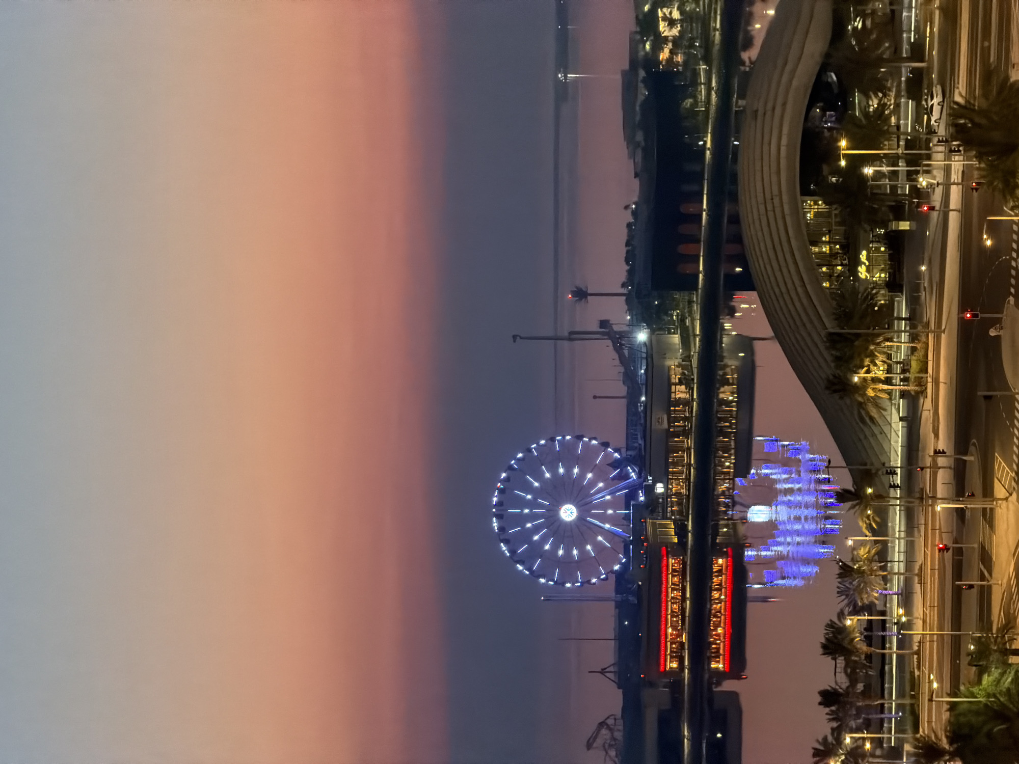 An aerial shot captures a waterfront cityscape at dusk, with a vibrant Ferris wheel illuminated by white lights against a gradient sky of pink, purple, and blue. The Ferris wheel is the dominant feature in the middle ground, reflecting on the still water below. To the left of the Ferris wheel, a building with a red illuminated facade is visible, also mirrored in the water with blue light reflections. In the foreground, a modern building with a distinctive arched roof is illuminated by yellow lights. Palm trees are scattered throughout the scene, lining a street with traffic lights, crosswalks, and a few parked cars. The water extends to the horizon, where a faint light from a distant structure can be seen. The overall scene depicts a lively urban waterfront area at twilight.
