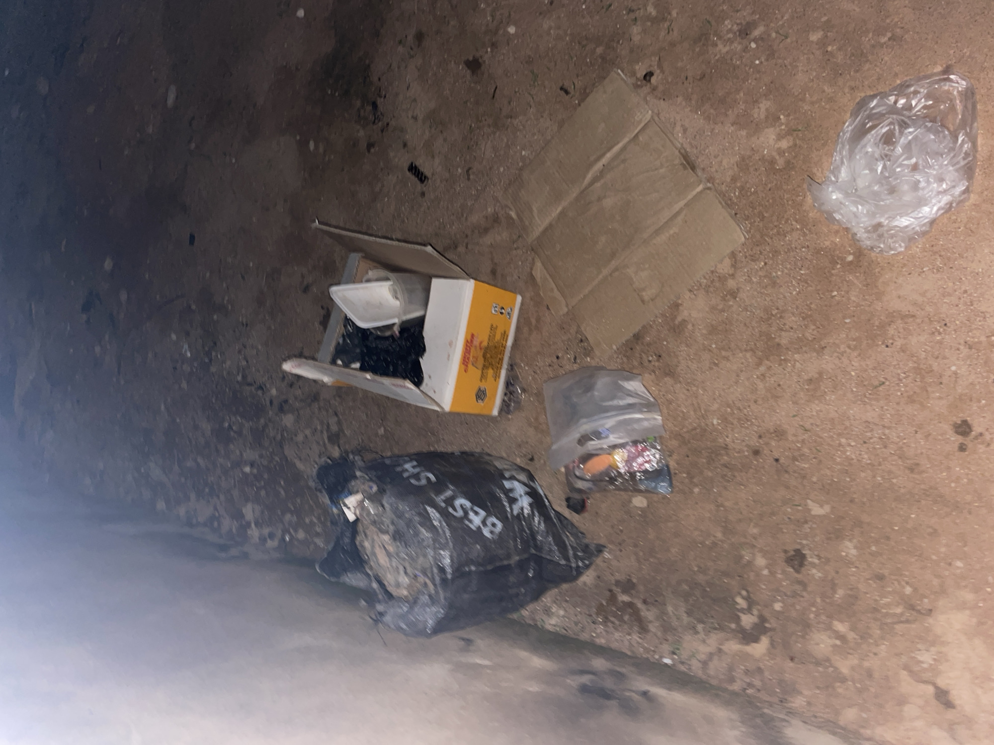 The image depicts an outdoor scene, possibly in an alleyway or undeveloped area, on a dirt or gravel surface. In the foreground, to the left, is a large, black plastic trash bag filled with debris, with the words "BEST SH" partially visible on its side. Next to it, slightly to the right and closer to the viewer, is a smaller, clear plastic bag containing various items, including what appears to be colorful wrappers or small objects. To the right of the trash bags, a cardboard box lies open, with some contents visible inside, including what looks like a white plastic container and dark material. Further to the right, a piece of folded brown cardboard lies on the ground. In the bottom right corner, a crumpled clear plastic bag is visible. The background shows a rough, textured wall on the left and a continuation of the ground surface. The lighting suggests it is either dusk, dawn, or an artificially lit area, with a strong light source in the upper left. The scene is located in Jalingo, Nigeria.