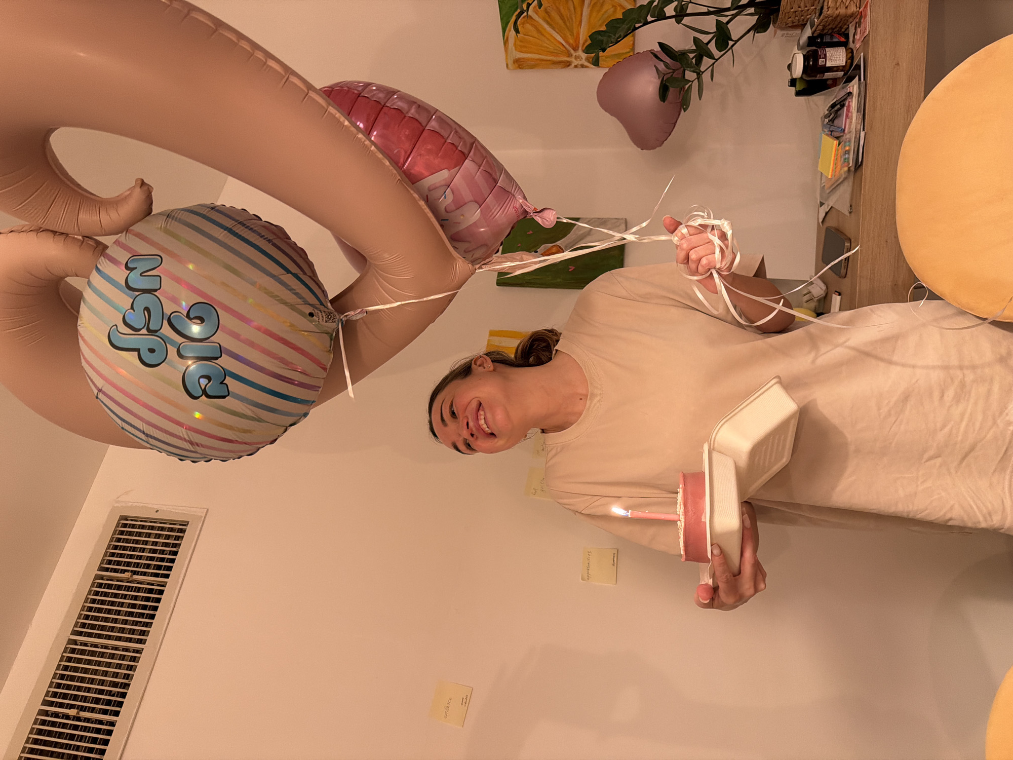 A woman stands inside a room holding a small cake with a lit candle, likely celebrating a birthday. She is surrounded by balloons, including large number balloons and a round balloon with the letters "BIG NSP" on it. The walls are cream-colored with several sticky notes attached to them. An air vent is visible in the lower-left corner. Other objects such as artwork, plants, and a shelf with items on it are visible in the background. The location is unknown.