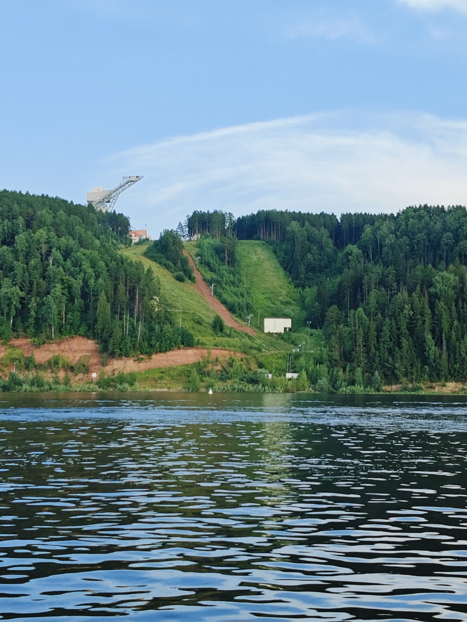 The image depicts a wide river in the foreground, with the opposite bank featuring a steep, forested hillside. At the top of the hill, a large, metallic ski jump structure is visible against a pale blue sky with wispy clouds. A cleared slope runs down the hillside, suggesting it is used for skiing or a similar activity. Several buildings are present on the hillside, including a prominent white rectangular structure. The water is calm with ripples and reflections, and a white buoy floats on the surface. The overall scene suggests a recreational or sporting area integrated with the natural landscape of Noviy, Russia.