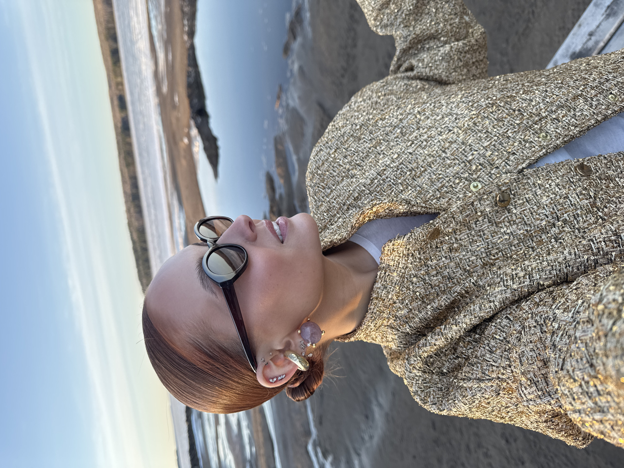 A woman poses for a selfie against a backdrop of water and beach in Chaikovsky, Russia. She wears sunglasses, multiple earrings, and a gold tweed jacket over a white top. The water is a light blue, and the beach appears sandy.