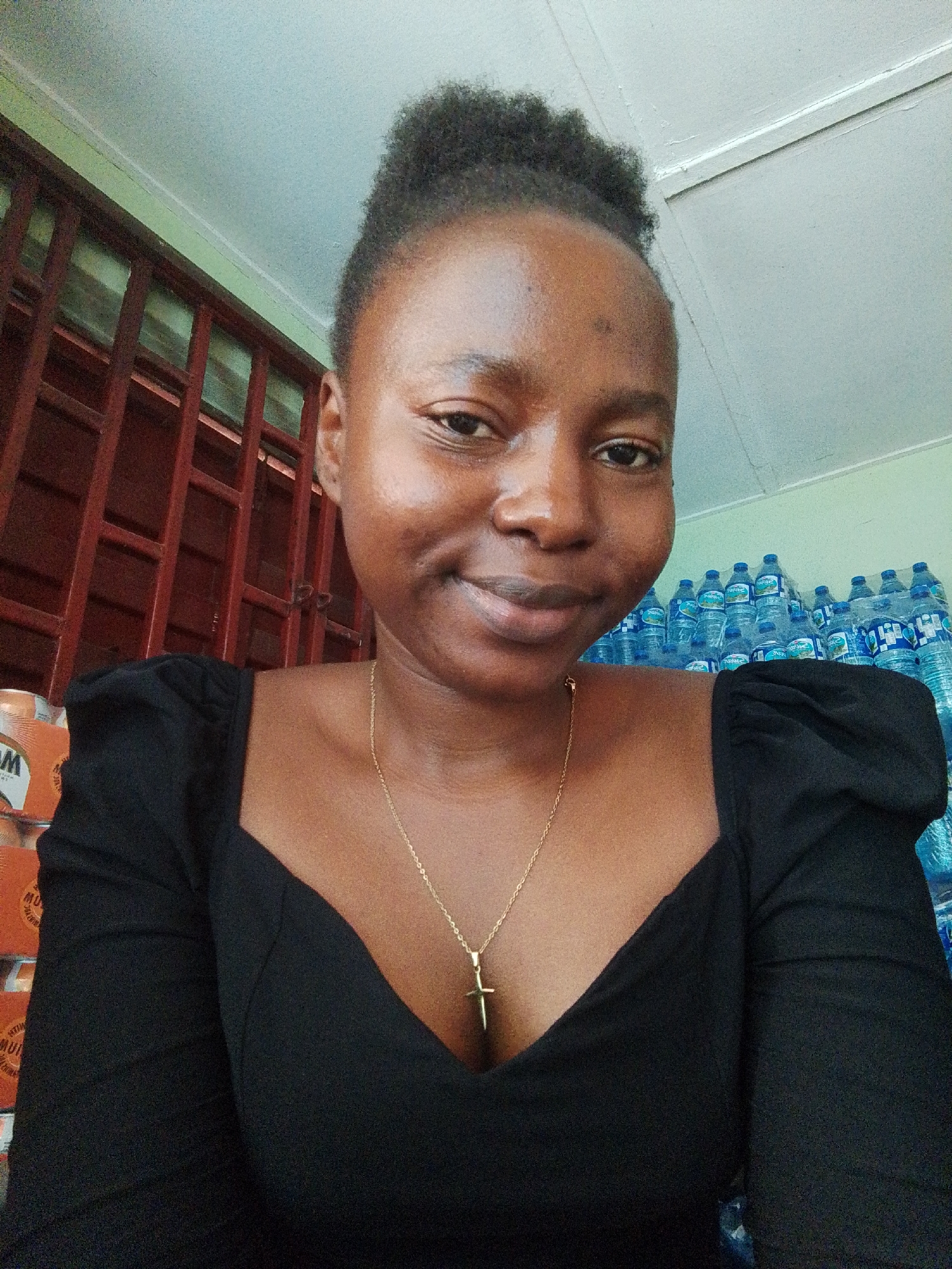 A medium-angle selfie taken in Jos, Nigeria, shows a young Black woman with dark hair styled upwards and slightly to the side. She is wearing a black top with puffed sleeves and a gold necklace with a cross pendant. Her skin has a sheen, reflecting the light. She is smiling gently at the camera.
Behind her, to the left, is a window with dark red grilles. To the right, stacks of bottled water are visible. Also visible to the left are stacks of cans. The background suggests a store or storage area. The ceiling is white. The lighting is adequate, but uneven.