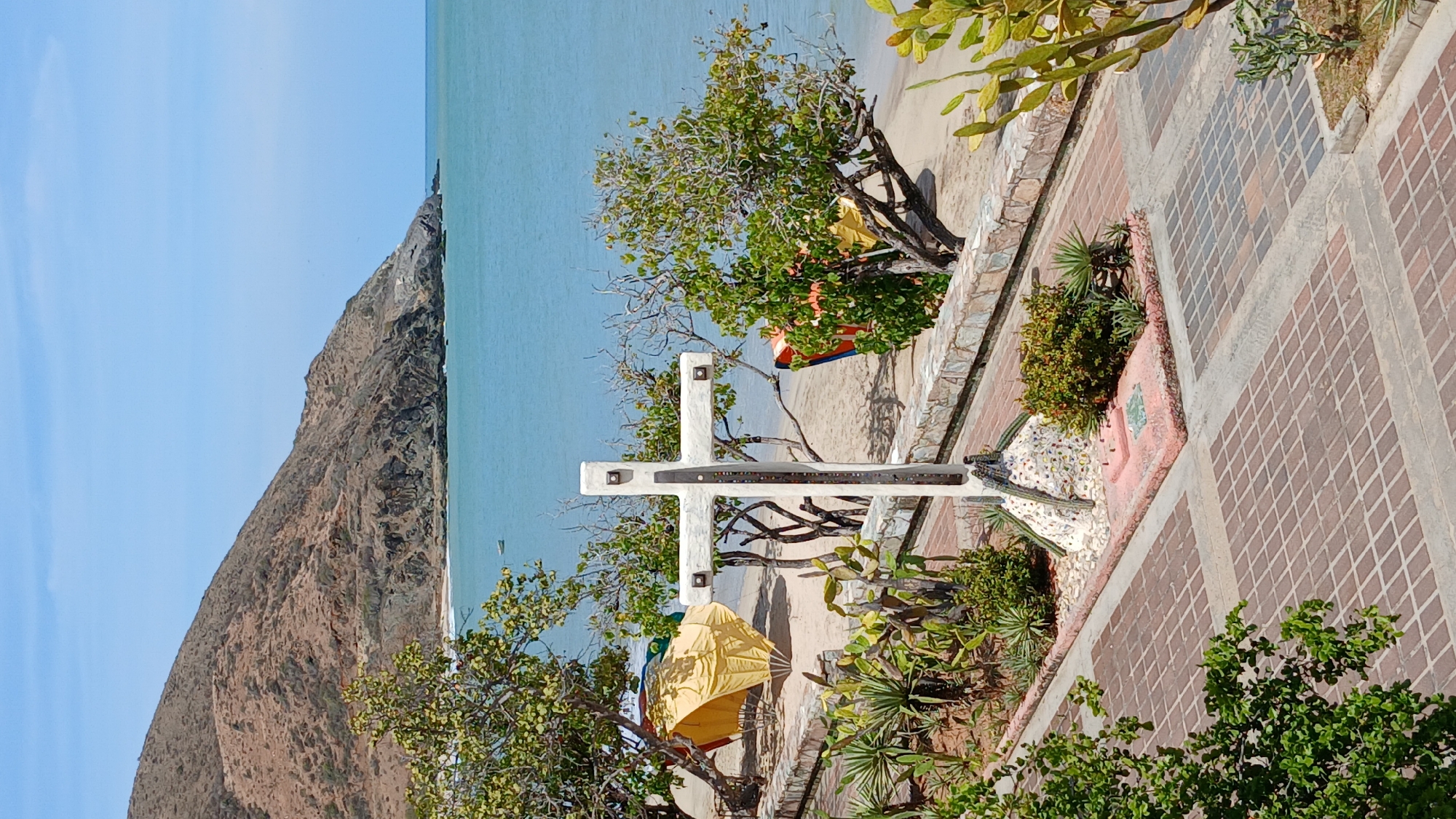 The image depicts a coastal scene in Pedro González, Venezuela, under a clear blue sky. In the foreground, a paved promenade composed of reddish-brown interlocking bricks is visible, flanked by garden beds containing various cacti, succulents, and small shrubs. A white concrete cross monument stands centrally within one of these planters, its base surrounded by a mosaic of colorful tiles and pebbles, and its main structure featuring embedded metallic squares. Behind the cross, a low stone wall separates the promenade from a sandy beach. On the beach, several yellow and orange tarpaulin structures, likely temporary shelters, are erected among scattered green trees with sparse foliage. The calm, light blue waters of the sea occupy the mid-background, with a single small boat visible offshore towards the left. A large, arid, rocky mountain dominates the left background, descending towards the coastline, which transitions to rugged rocky outcrops on the right. No individuals are present in the scene.
