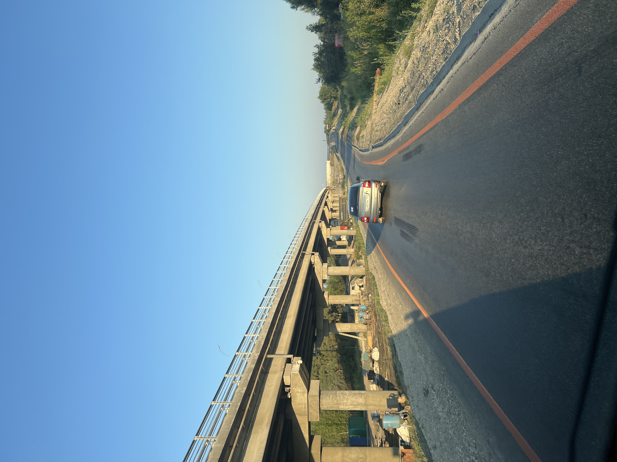 The image depicts a road with a car driving under an elevated highway that is under construction. The scene appears to be in Katkovskii, Russia, given the context. The highway is made of concrete and supported by pillars, with metal railings along its edge. Construction materials and equipment are visible beneath the highway, including what looks like a sandblaster, containers, and bags of materials. The road itself is asphalt, with orange lane markings. The surrounding environment is a mix of grassy areas, trees, and shrubs. The sky is clear and blue, indicating daytime with clear weather. The perspective suggests the photo was taken from a vehicle, likely a car, moving along the road.