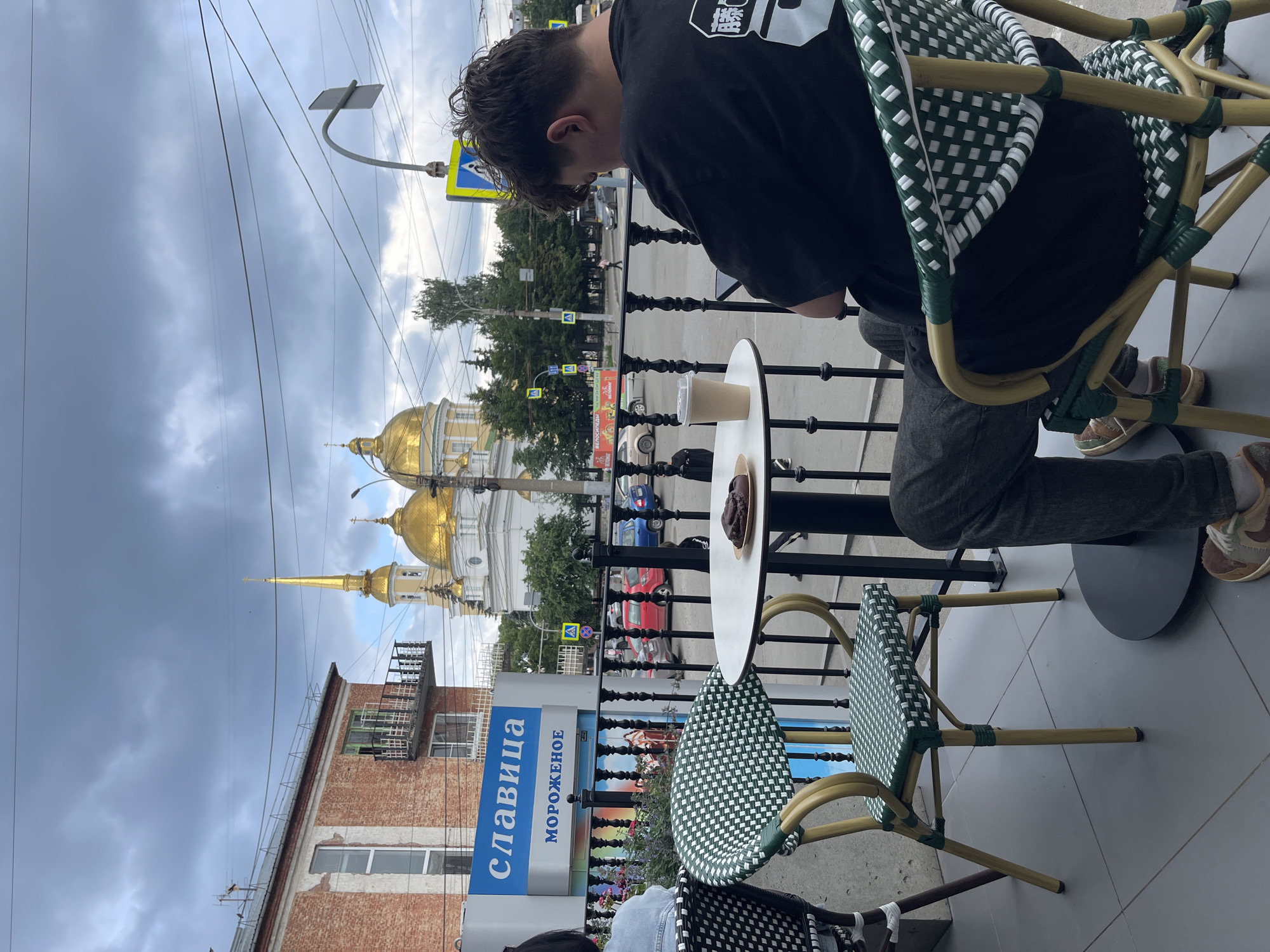 The image shows a street-level view from a cafe patio in Izhevsk, Russia, with a view of the Ascension Cathedral. In the foreground, a person sits at a small round table with a pastry and a cup of coffee, facing away from the camera and looking at their phone. Two cafe chairs are visible, one occupied by the person and another empty. Behind the patio, a street with cars and pedestrians is visible, and further in the background, the golden domes of the Ascension Cathedral are prominent against a cloudy sky. A building with a sign that reads "СЛАВИЦА МОРОЖЕНОЕ" (Slavitsa Ice Cream) is to the left of the cathedral. Traffic lights and road signs are also visible.