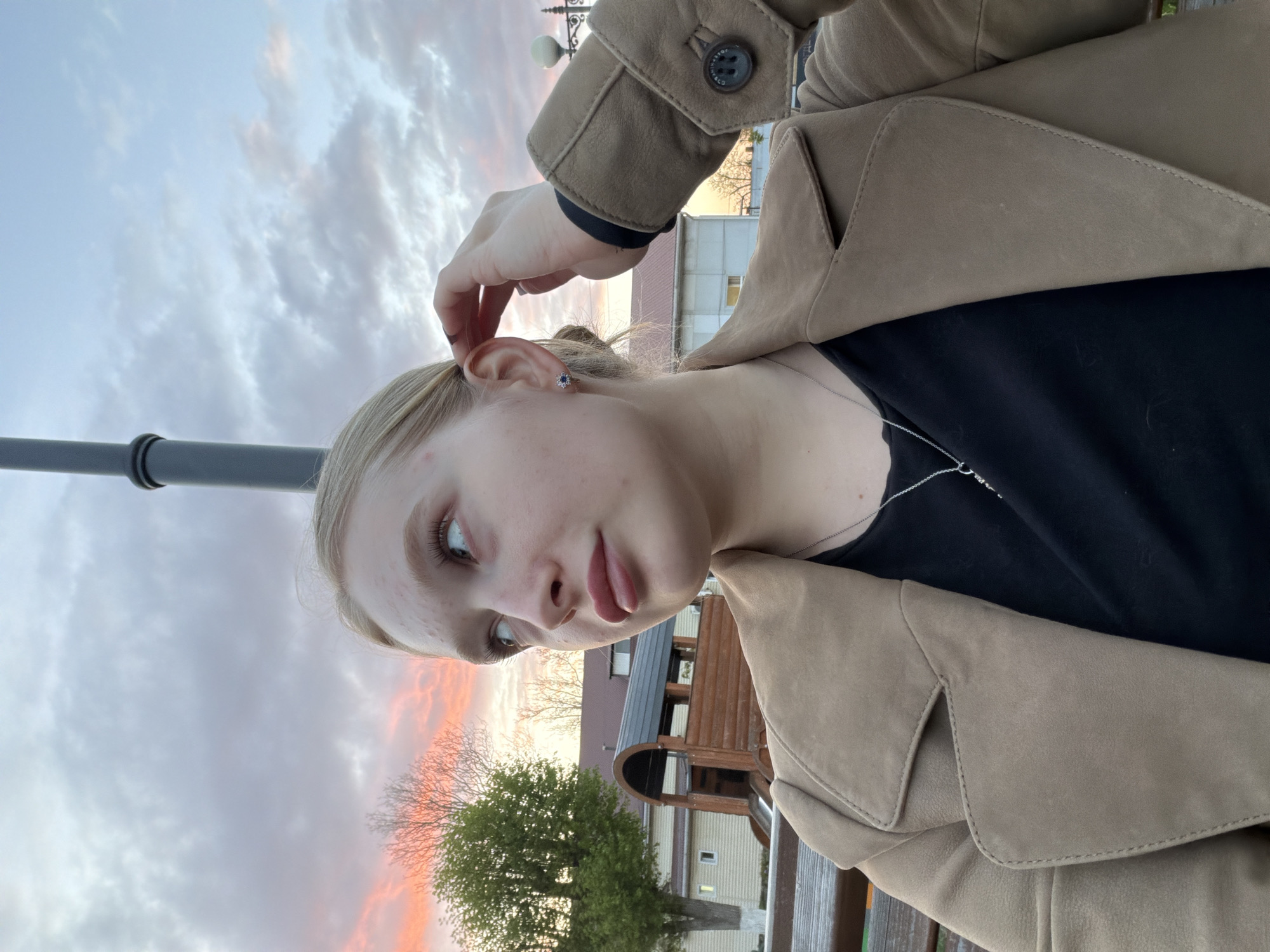A young woman with blonde hair is looking at the camera. She is wearing a black shirt and a tan jacket. Her right hand is touching her hair near her ear. The sky is cloudy with a pink and orange sunset. There is a streetlight pole visible. In the background, there is a building that appears to be made of wood, suggesting the photo was taken in the city of Yuzhny, Russia, given the context. A green tree is also visible in the lower center of the image.