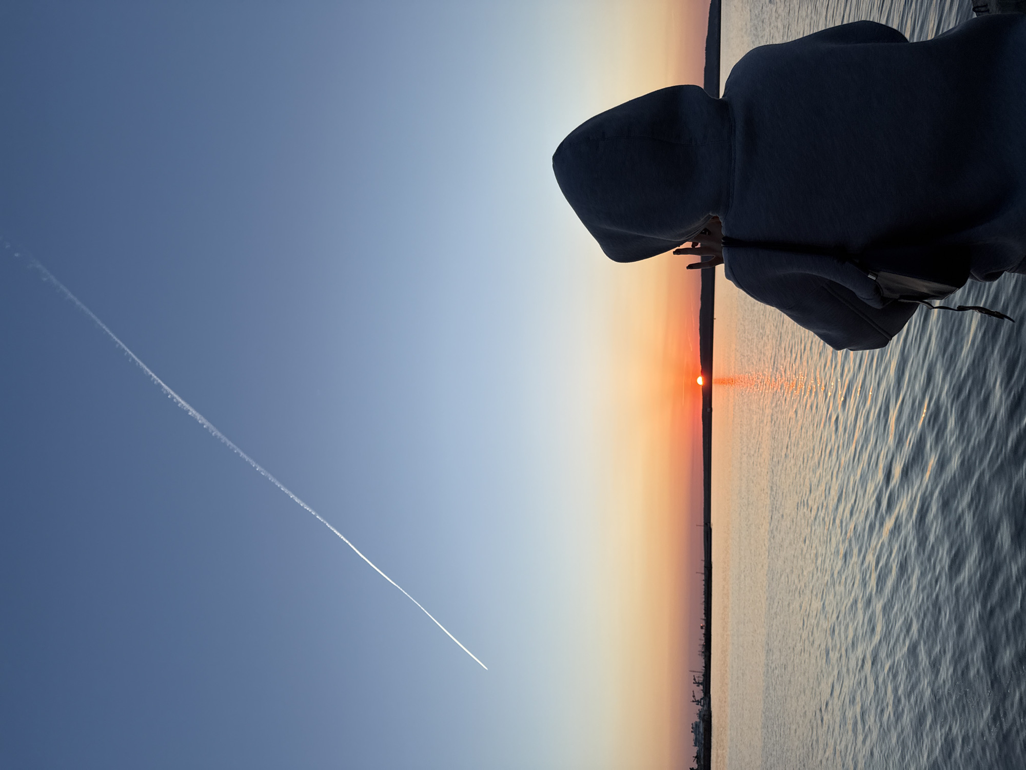 The image shows a sunset over a body of water. In the upper right of the image is a person in a grey hooded sweatshirt. The sky transitions from dark blue to orange and the sun is just above the horizon. A contrail crosses the sky diagonally. On the horizon, on the water's edge, there is a cityscape silhouette. This scene is in the city of Chaikovsky, Russia.